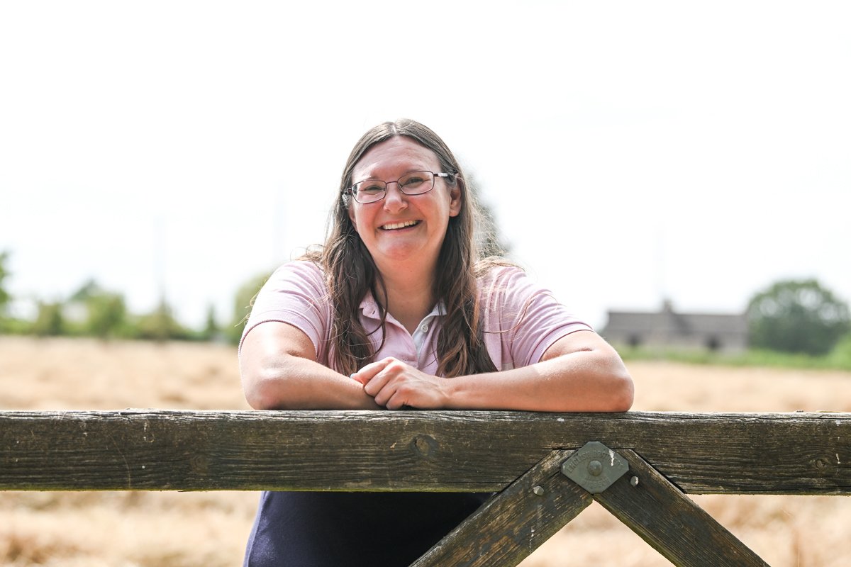 A woman with long brown hair, glasses, and a pink shirt smiling and resting her arms on a wooden fence outdoors on a sunny day.
