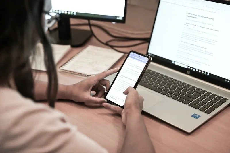 Person holding a smartphone in front of a laptop and two monitors on a desk, with notes beside them.