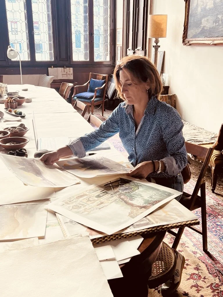 A woman sitting at a large dining table with art prints and papers spread out in front of her, in a room with stained glass windows, a side table with a lamp, and framed artwork on the wall.