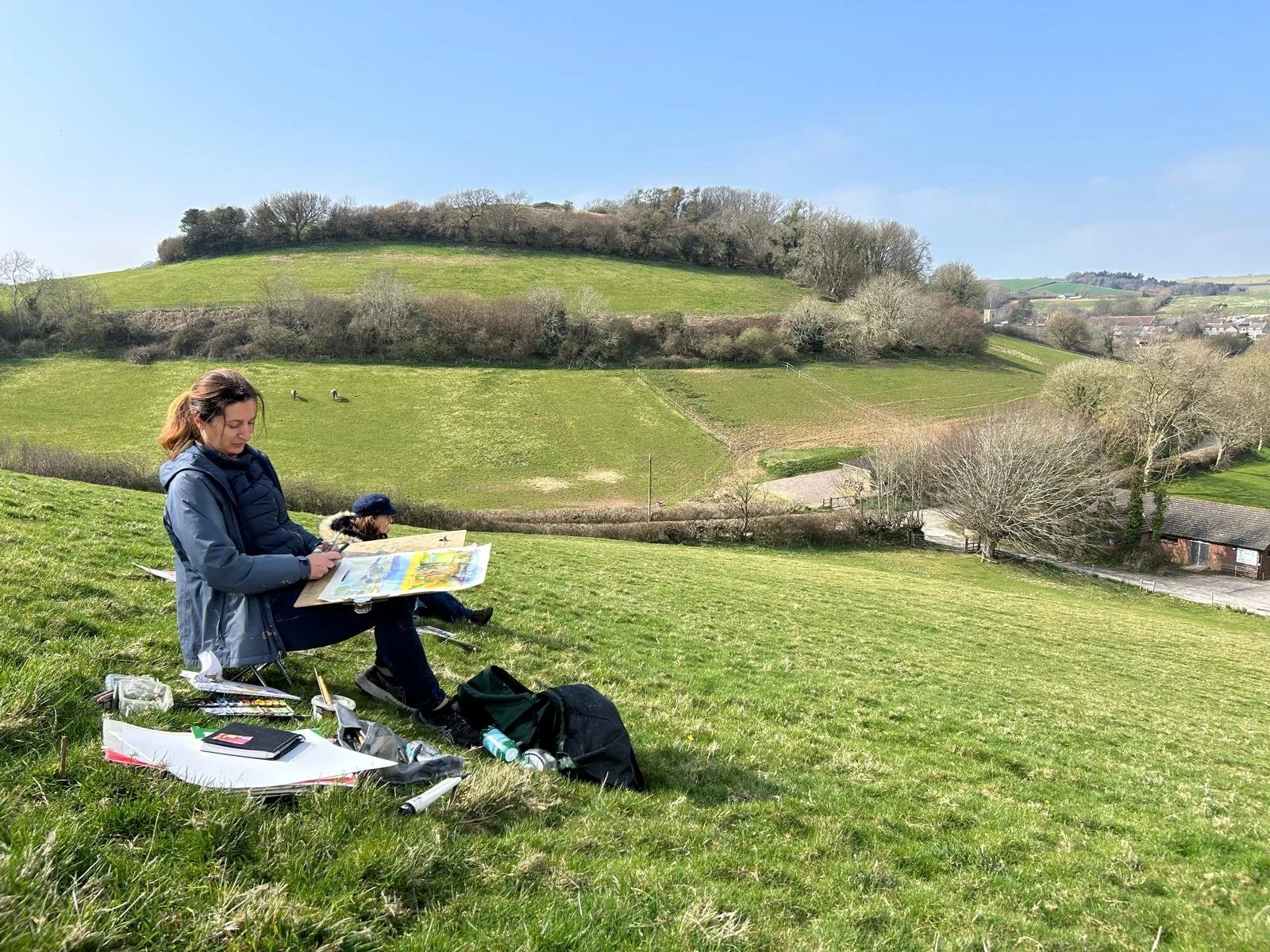 Two women sitting on a grassy hillside, one looking at a large colorful map, with art supplies and notebooks nearby, overlooking a scenic rural landscape with rolling hills, trees, and a distant small town.