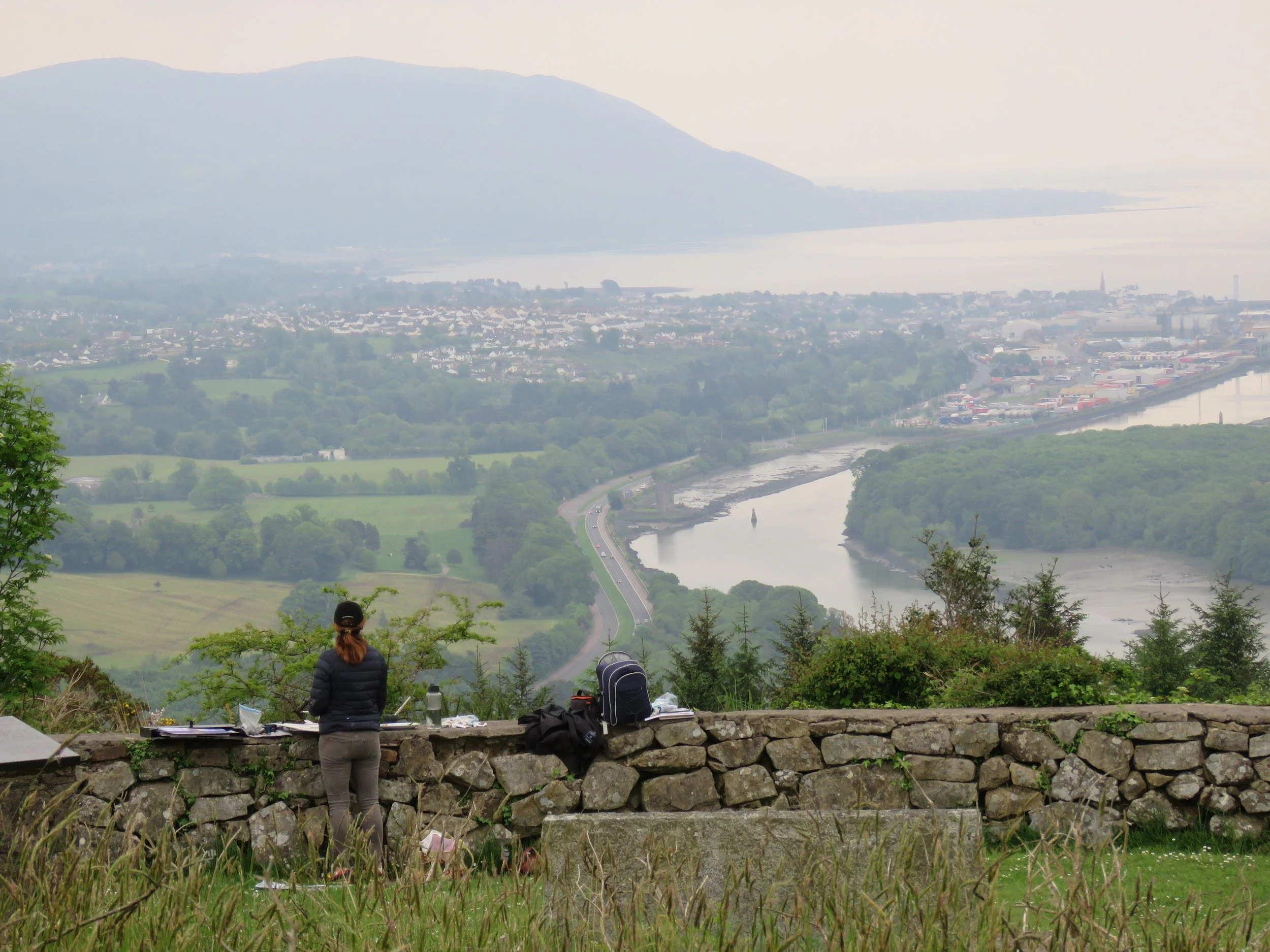 A person standing next to a camcorder and a backpack on a stone wall, overlooking a landscape with a river, a town, and mountains in the background.