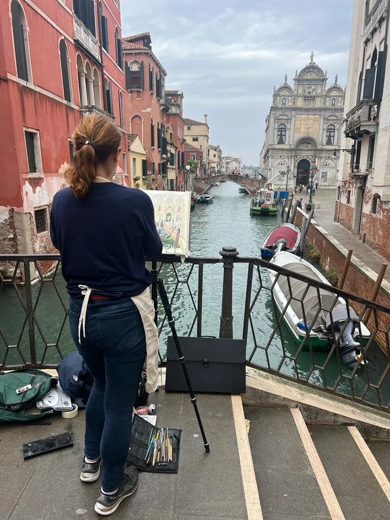 A woman painting on an easel by a canal in Venice, Italy. She has art supplies on the ground and is standing on a small bridge with buildings and a church in the background.