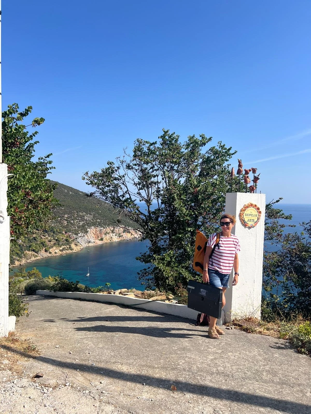 A woman with sunglasses and a striped shirt stands smiling near a white post with a circular sign that reads 'Shopelos Arts' with decorative flowers. She has a backpack and a briefcase. There is a scenic coastal view with a cliff, water, and a sailbo