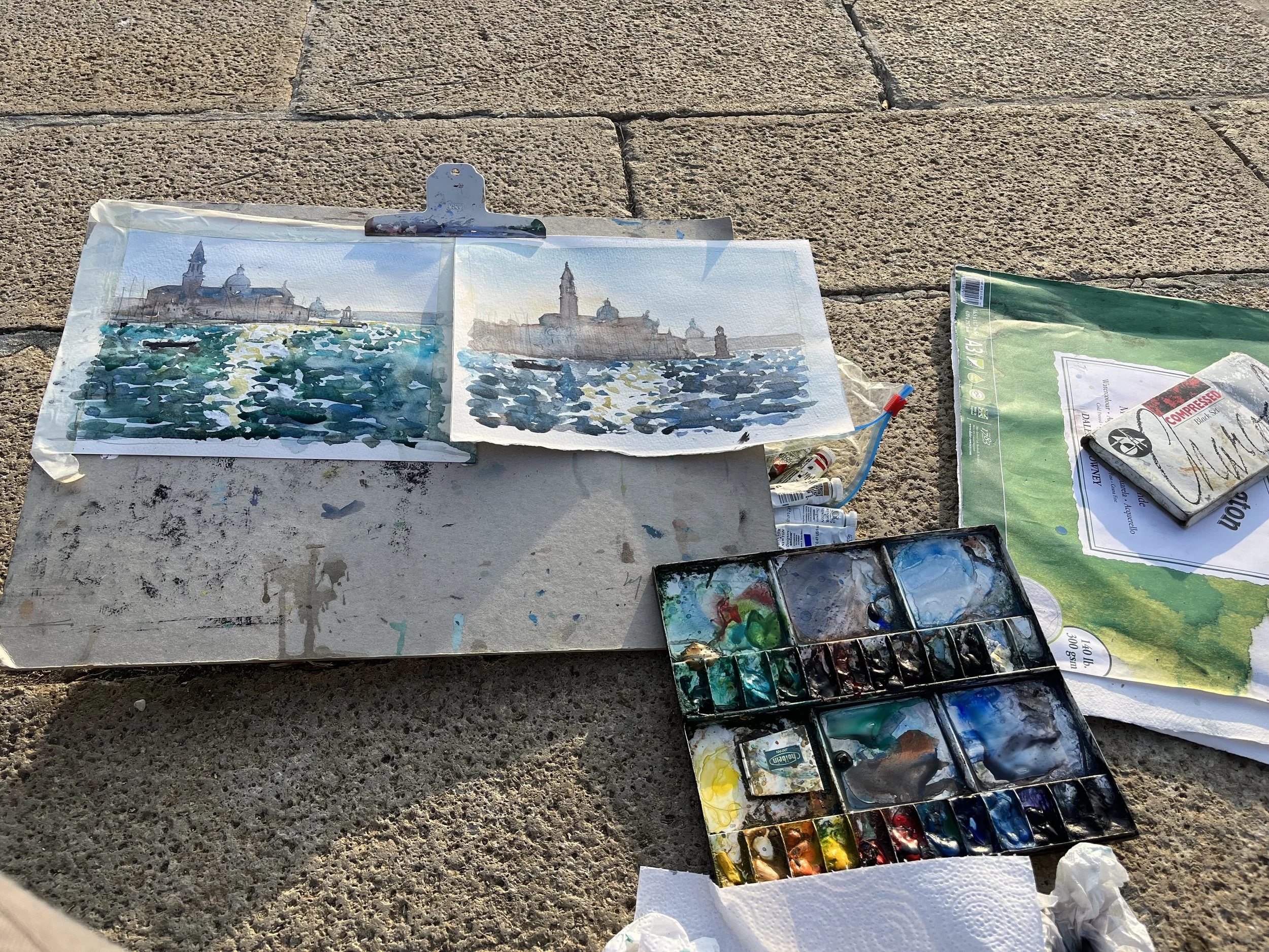 Paintings of Venice on paper with watercolor paints, watercolor palette, and art supplies on a rough outdoor surface.