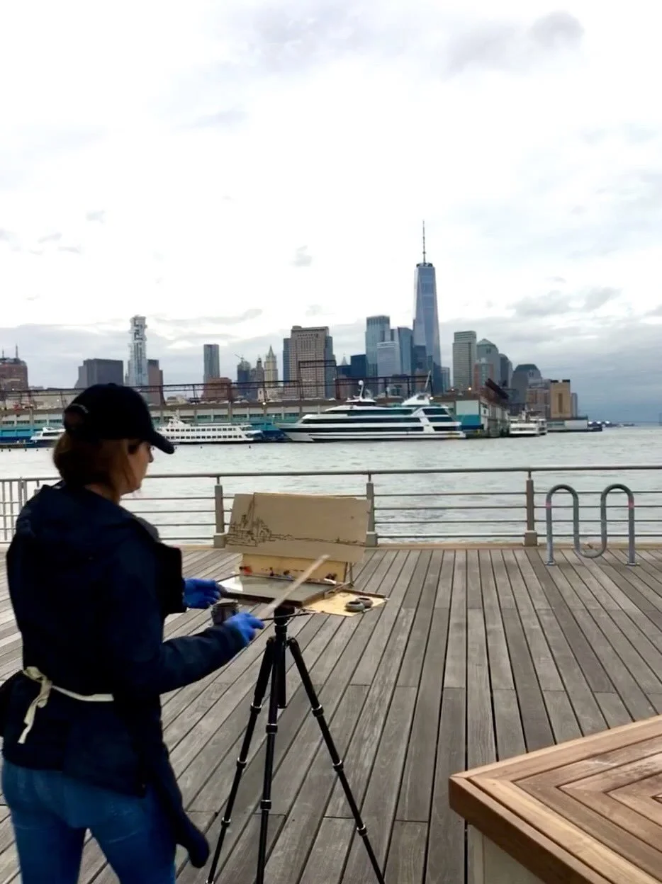 A person painting on a canvas set up on an easel on a pier with the New York City skyline, including the One World Trade Center, in the background.