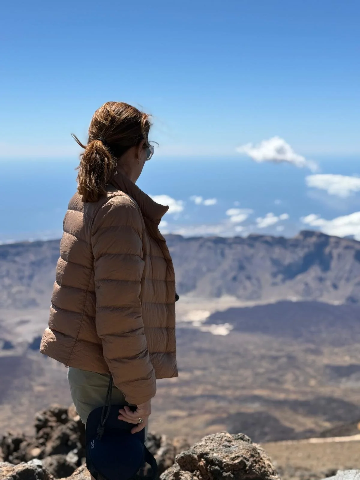 A woman standing on rocky terrain, facing a mountainous landscape with volcanoes, ocean in the background, and a blue sky with clouds.