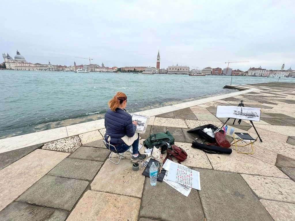 A woman painting on a small chair by the water in Venice, Italy, with her art supplies and a sketch of a ship on an easel nearby.