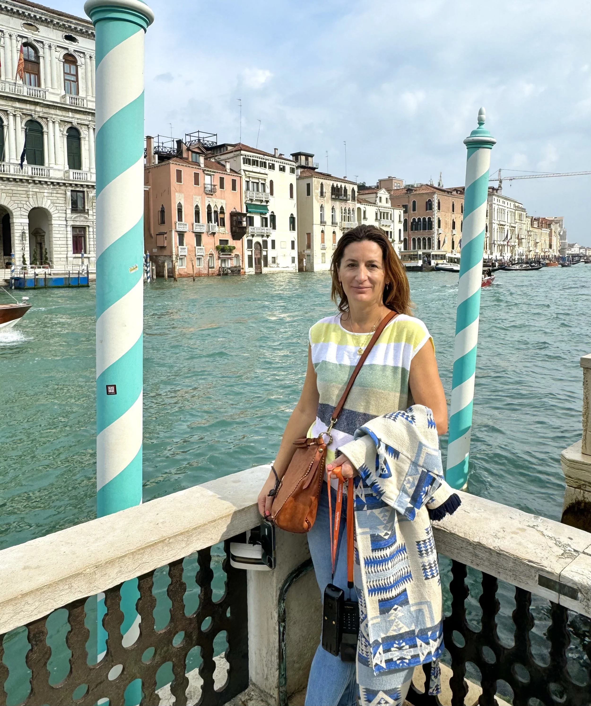 A woman standing on a stone bridge over a canal in Venice, Italy, with colorful historic buildings in the background, two striped teal and white poles in front, and boats on the water.