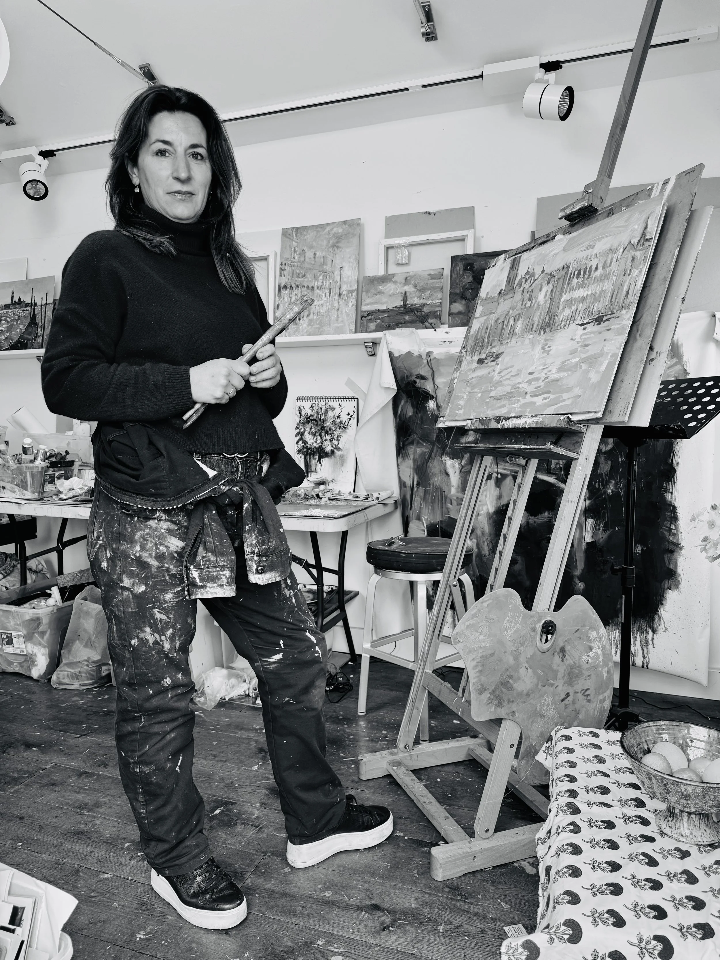 A woman with dark hair, wearing a black turtleneck sweater and paint-splattered pants, stands in an art studio holding a paintbrush. She is in front of an easel with a painting of a city by the water, surrounded by various paintings, art supplies, and a table with a bowl of eggs and a patterned cloth.