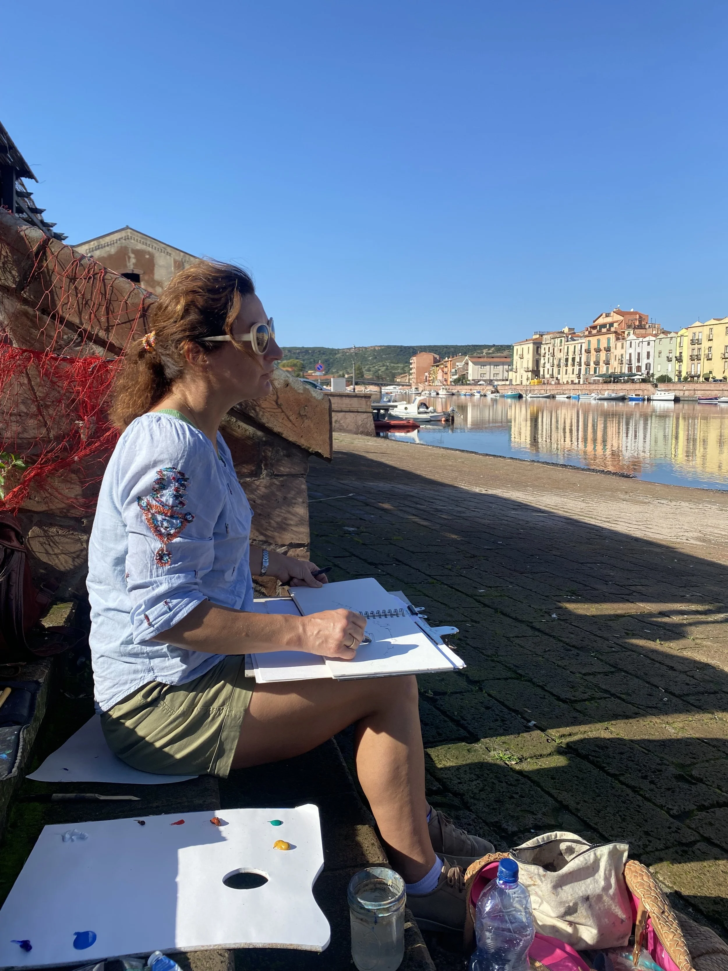 A woman sitting on a bench by a waterfront, sketching or painting in a sketchbook. She is wearing sunglasses, a white embroidered blouse, beige shorts, and sneakers. She has a backpack, a water bottle, and painting supplies nearby. The background fea