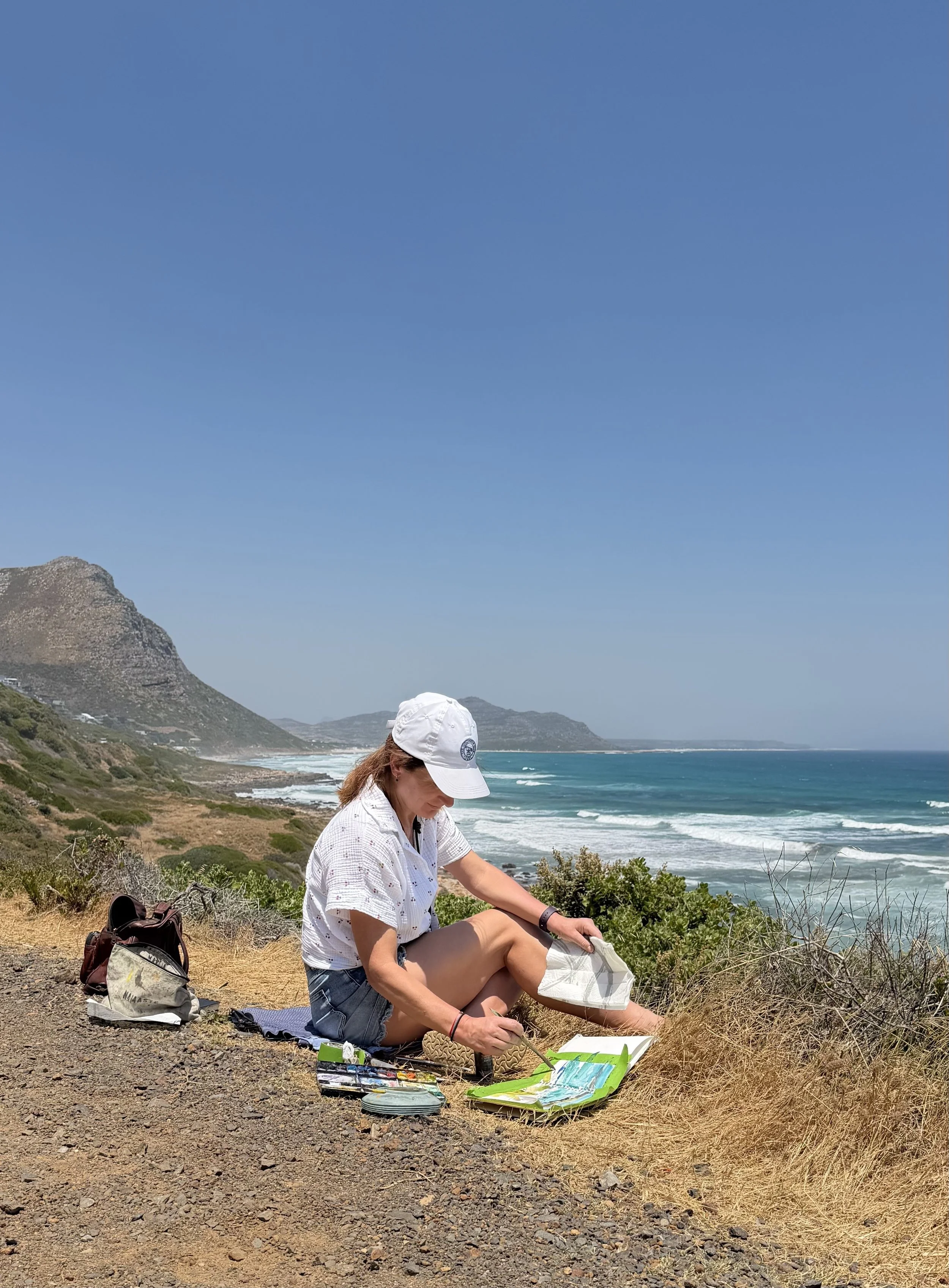 A woman sitting on a rocky coastal path with the ocean and mountains in the background, painting on a canvas. She wears a white cap and casual clothes, with art supplies and a backpack nearby.