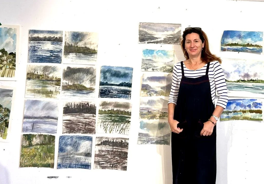 Woman standing in front of a wall displaying various landscape watercolor paintings.