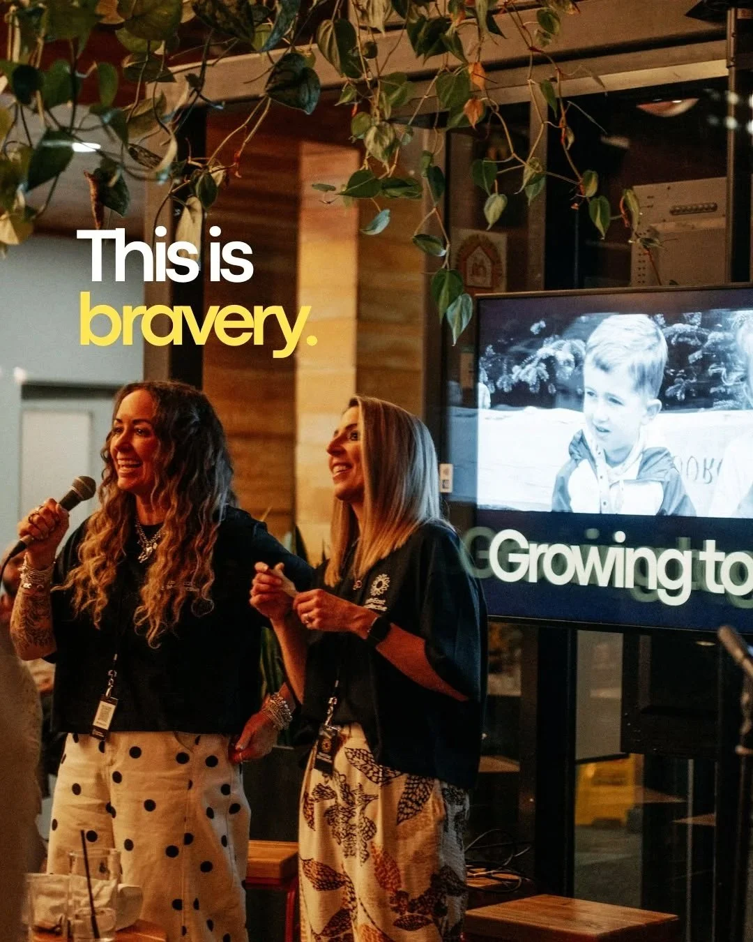 Two women standing together indoors, smiling and holding hands while one speaks into a microphone; a large screen in the background shows a black and white photo of a young boy, with the text 'Growing to'. The scene is warm and celebratory, with hanging greenery overhead.