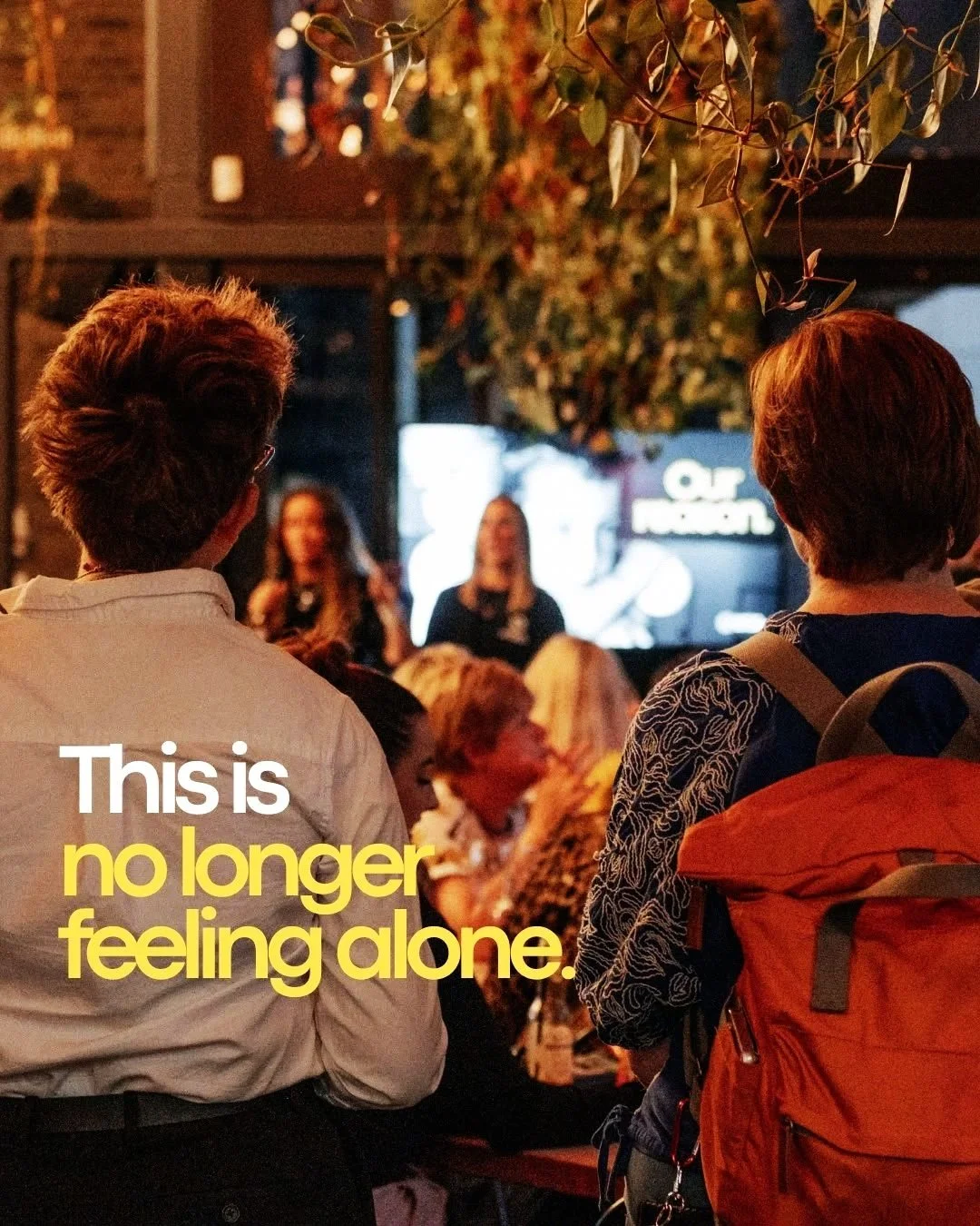 People gathered at a live event in a dimly lit venue with a stage and a screen in the background. The photo shows the backs of two individuals, one with brown hair wearing a white shirt and the other with short hair carrying an orange backpack. The scene includes an audience sitting and standing, with some focusing on the stage. Overlaid text reads, 'This is no longer feeling alone.'