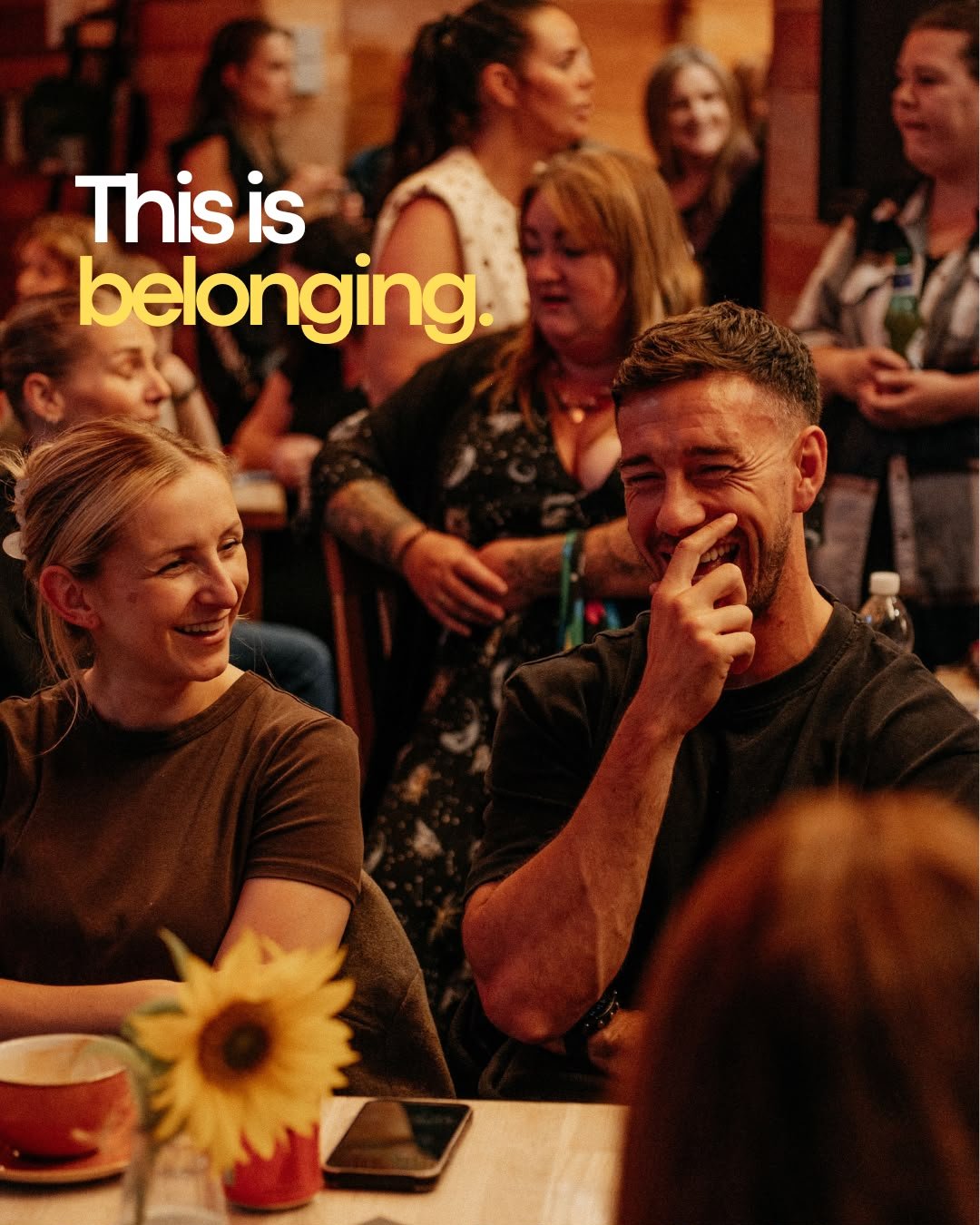 People sitting together, smiling, and laughing in a cozy, warmly lit setting that looks like a restaurant or cafe.