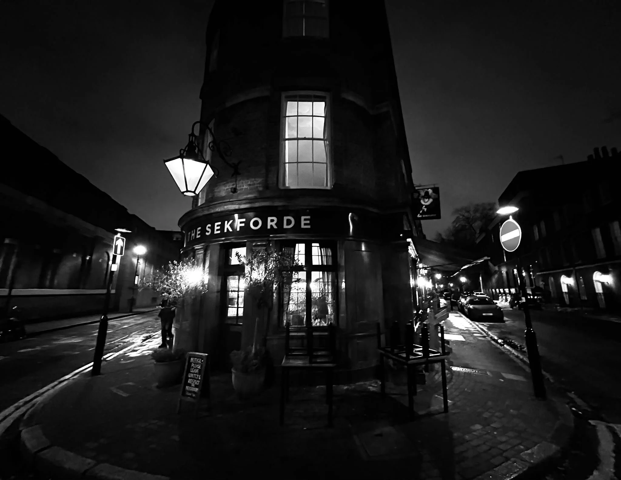 Night view of a street corner with a round building labeled "THE SEKFORDE," illuminated windows, a street lamp, a parking sign, and a man walking on the sidewalk.