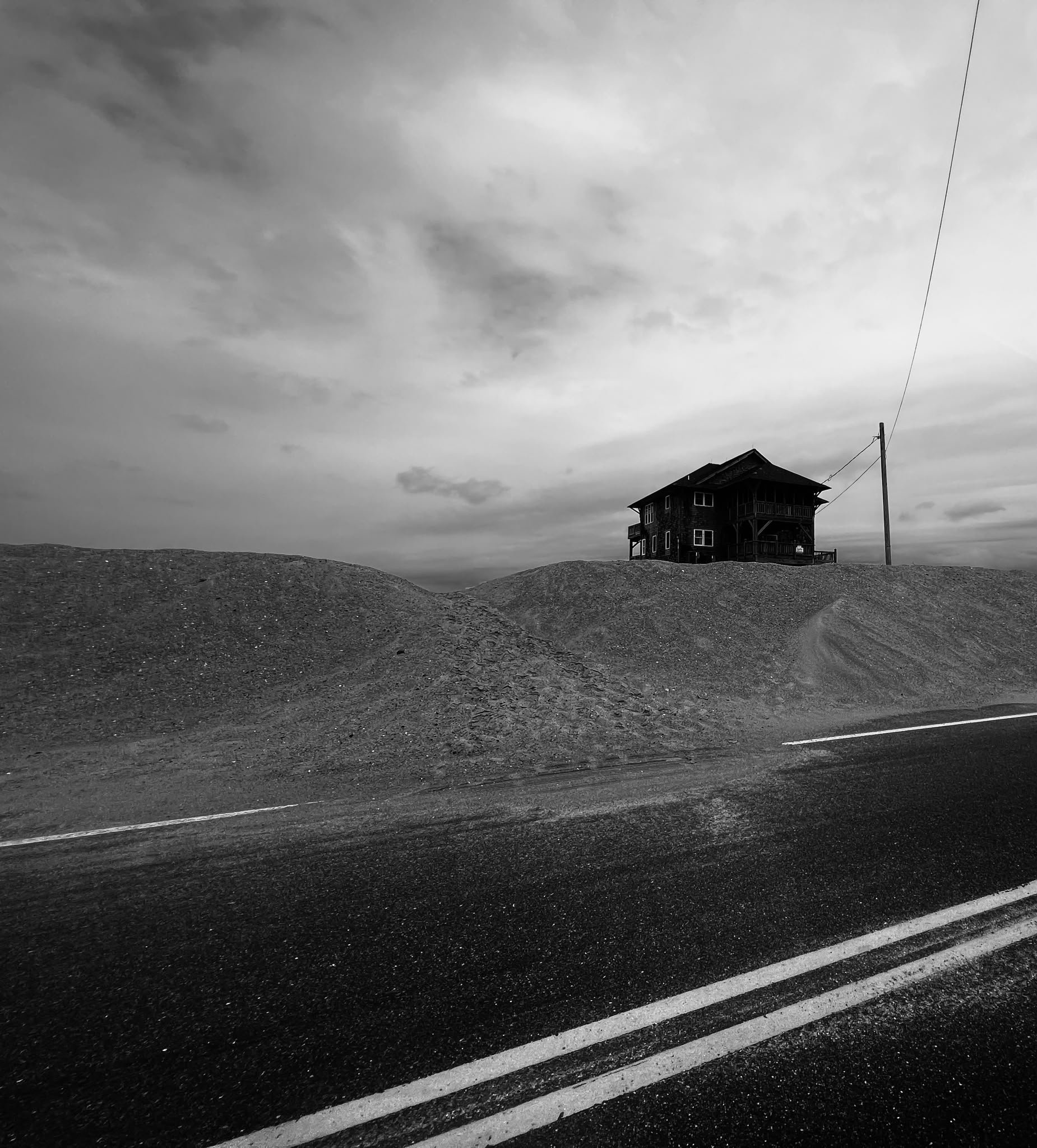 Black and white photo of an empty road with a shell of a house on a hill, cloudy sky, and power lines.