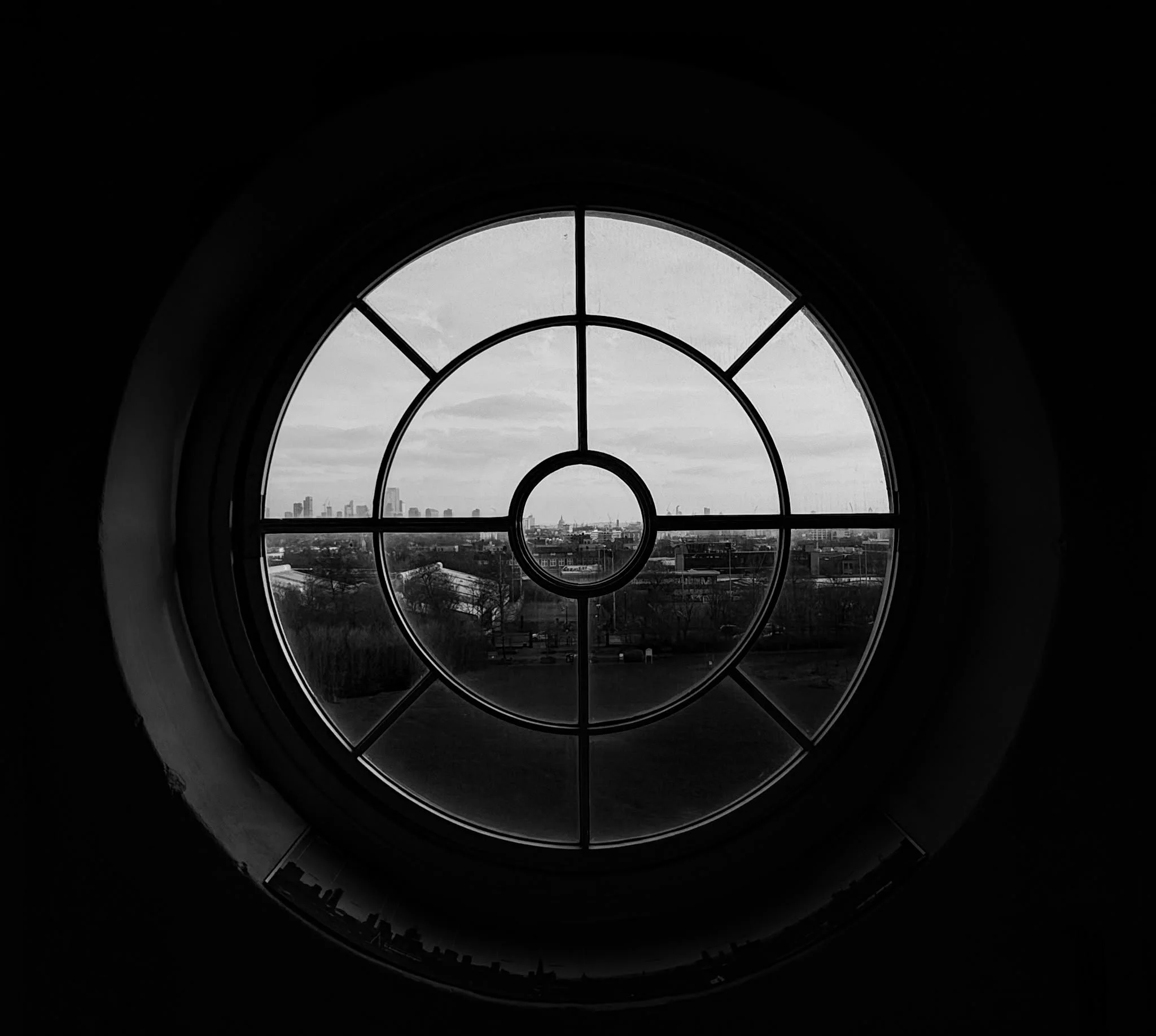 A large circular stained glass window with a view of a city skyline in the distance, taken from inside a dark room.