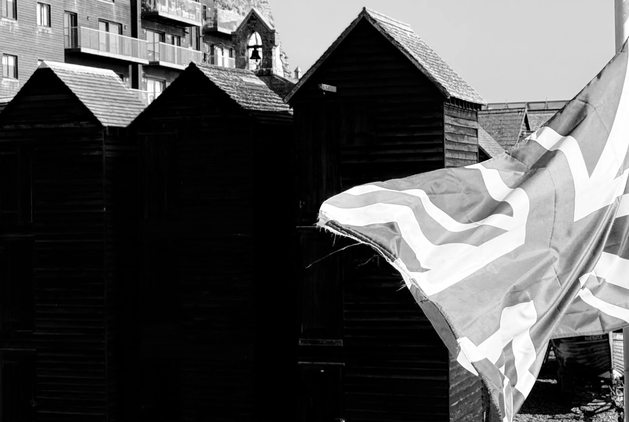 Black and white photo of a small wooden house with a sloped roof, set against a background of modern apartment buildings. A flag with a partial Union Jack design is in the foreground.
