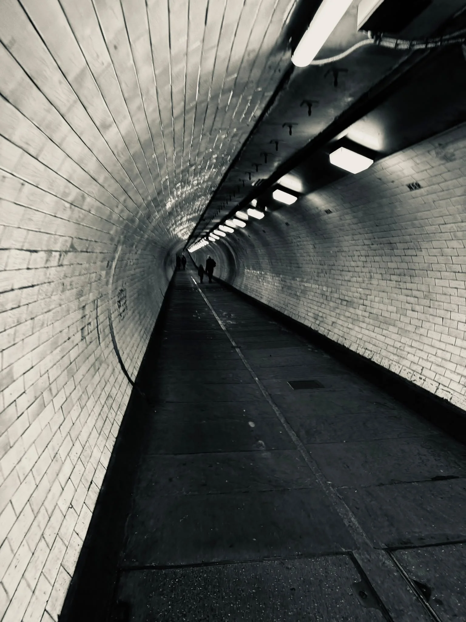Inside a tunnel with brick walls, fluorescent lights, and a few people walking in the distance.