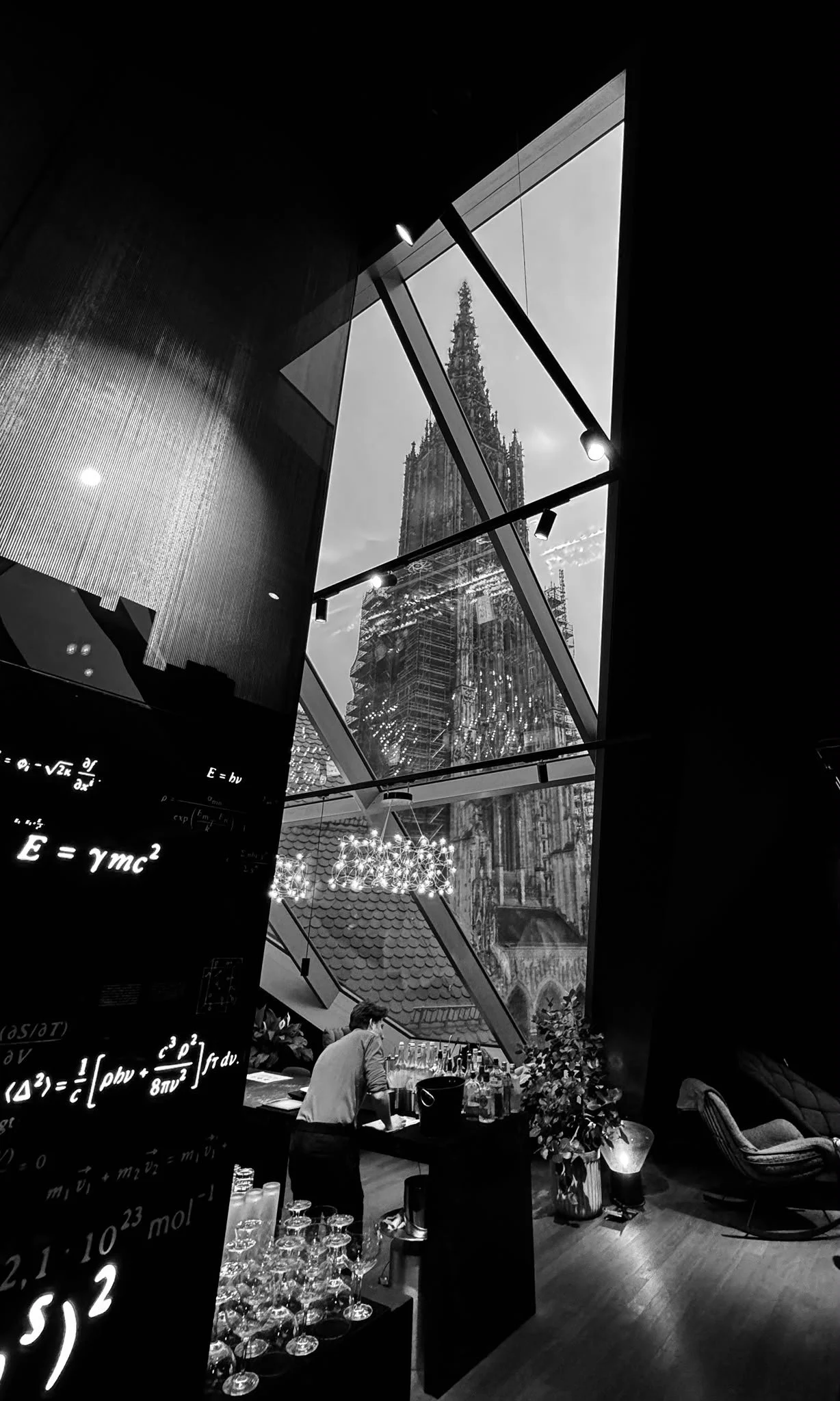 Black and white photo of an indoor space with large windows showing a cathedral and a tall tree outside. Inside, there are tables, a bar area, a person preparing drinks, and decorative lighting. A digital screen displays scientific equations.