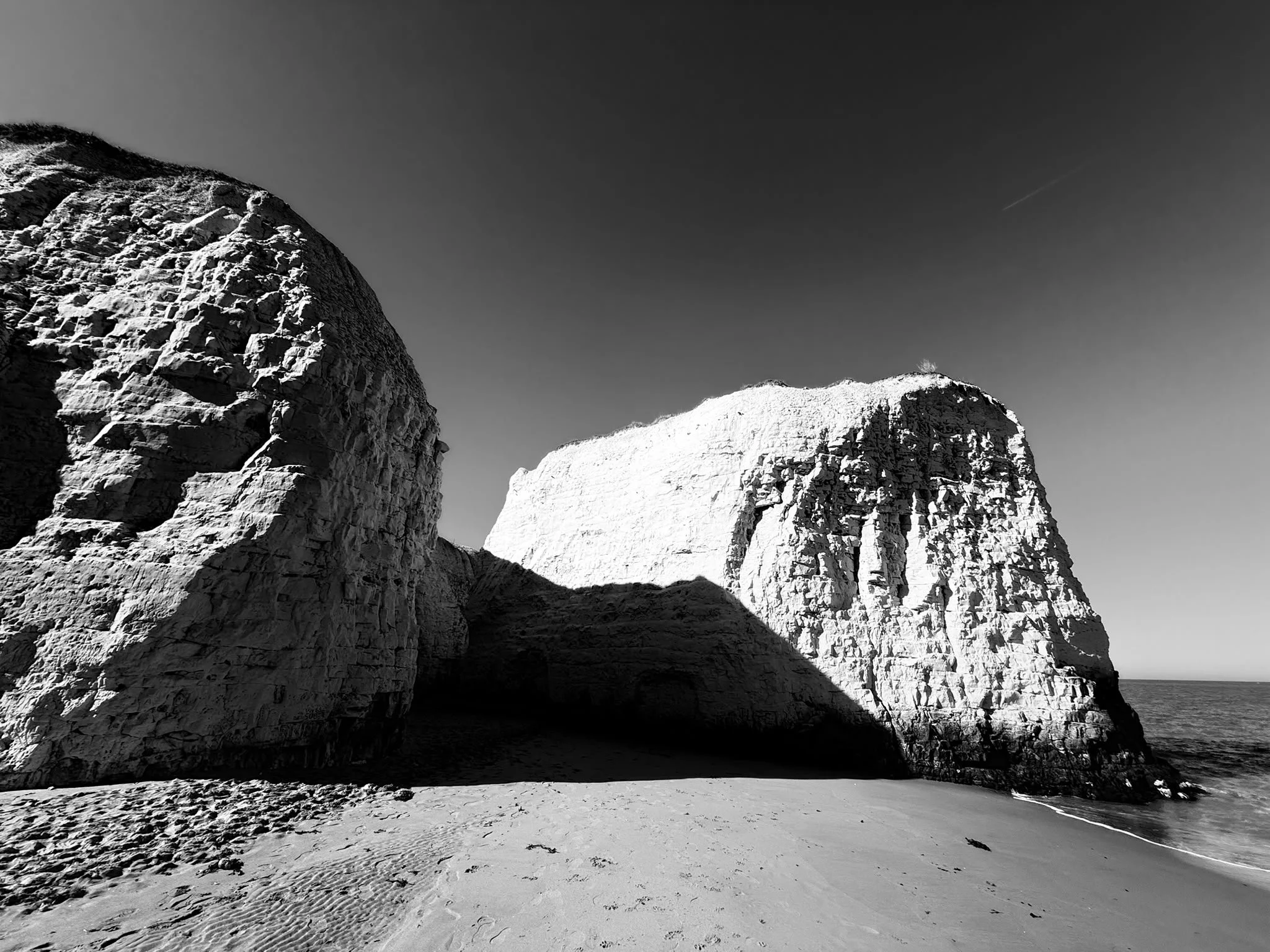 Black and white photo of large rocky cliffs near a sandy beach with ocean in the background.
