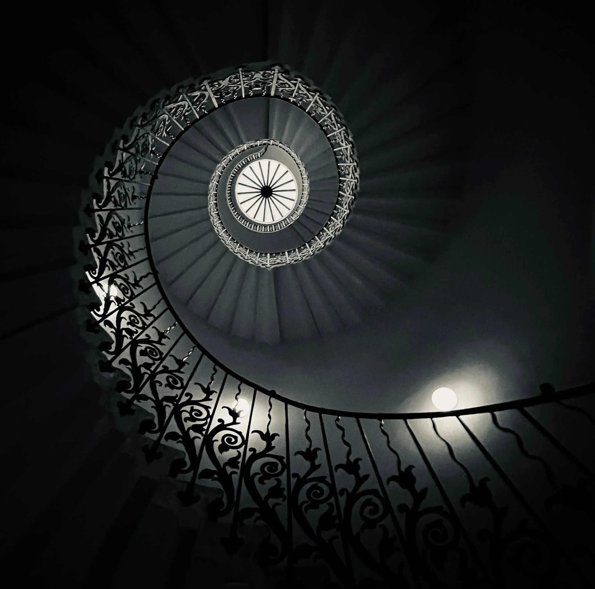 Spiral staircase viewed from below with ornate black wrought-iron railing and a central light fixture at the top.