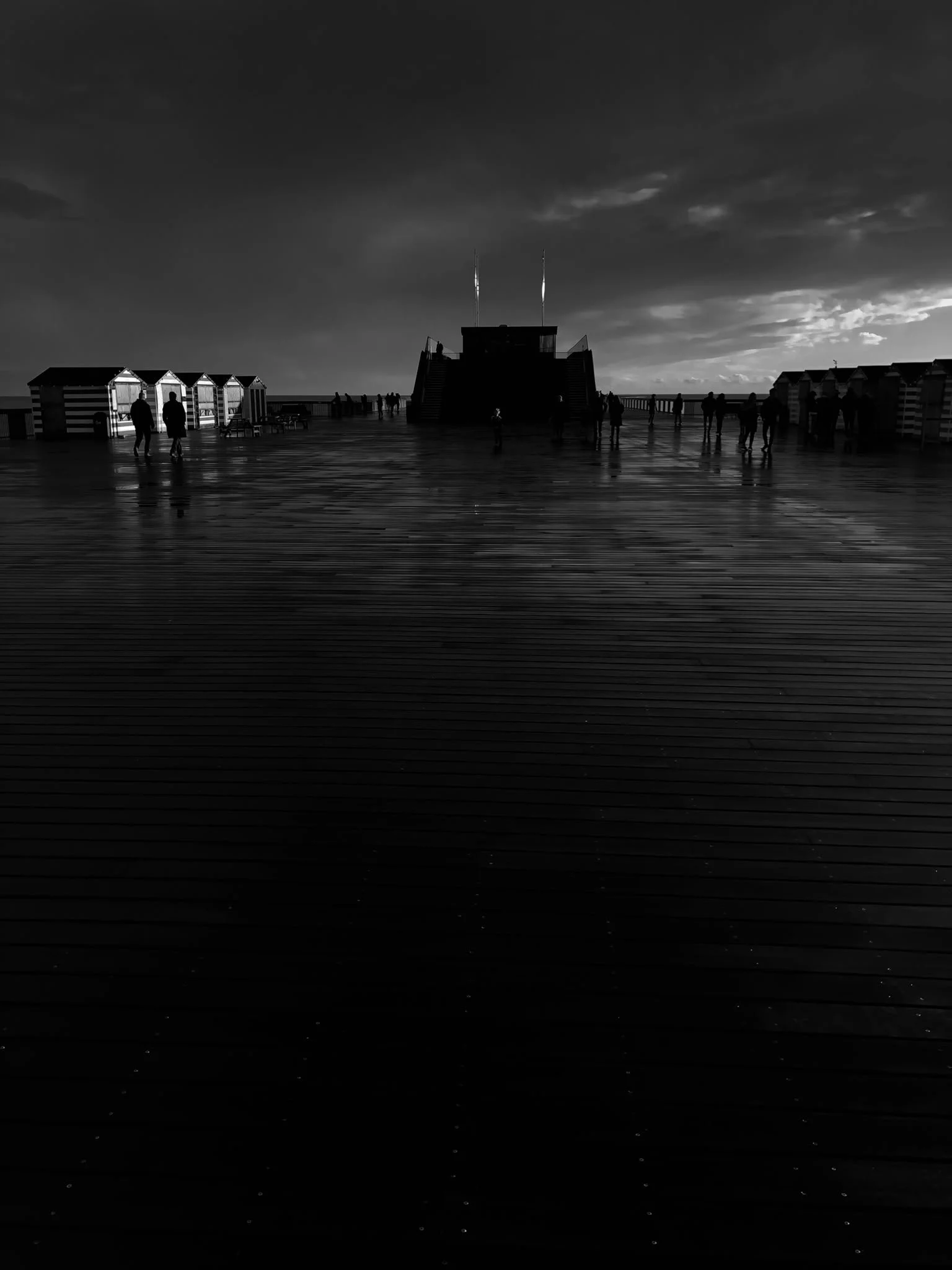 Silhouettes of people walking on a dark, wet boardwalk at sunset, with striped beach huts and a raised platform in the background.