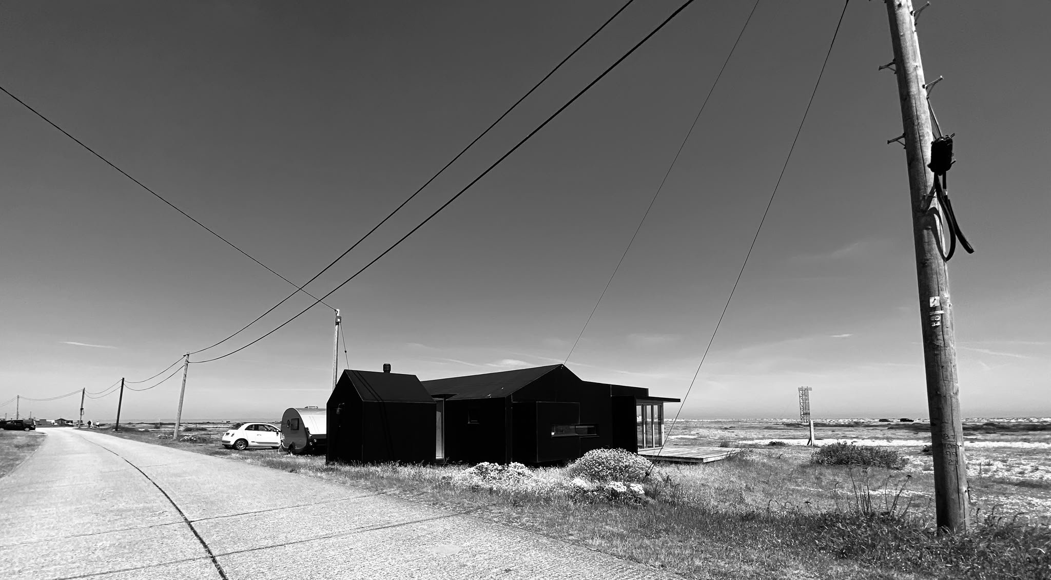 Black and white photo of a rural landscape with utility poles, power lines, a small house, and cars parked nearby under a clear sky.