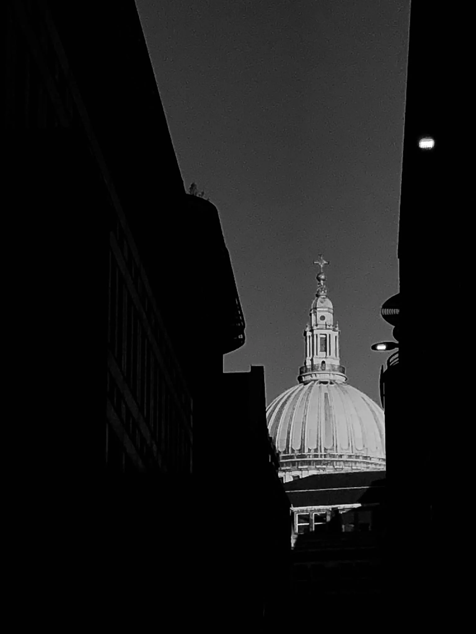 Black and white photo of a church steeple and dome seen between dark silhouettes of buildings at night.
