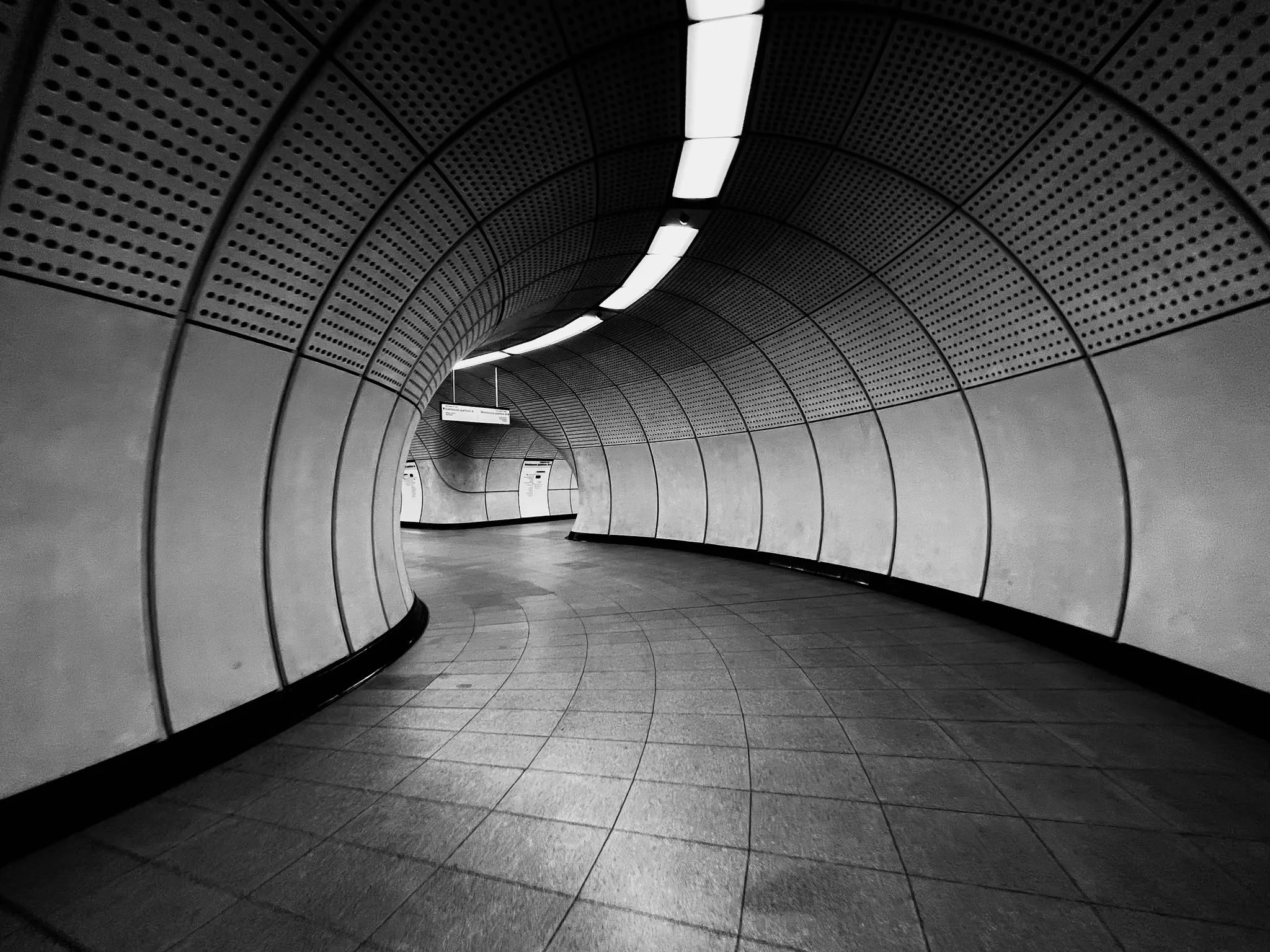 Underground subway tunnel with curved walls and ceiling, illuminated by linear fluorescent lights.