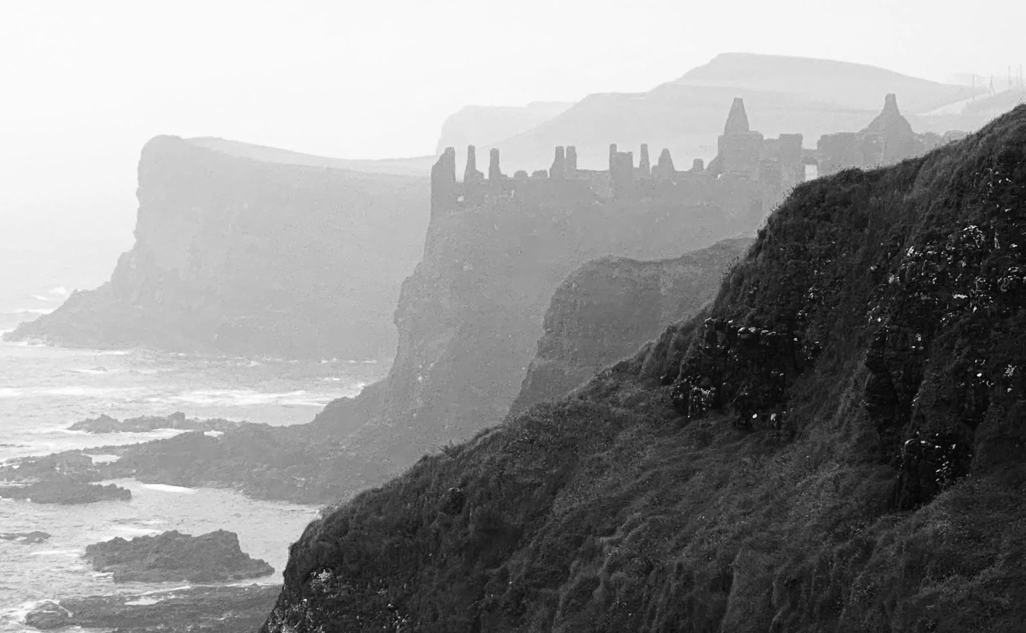 Black and white photograph of dramatic coastal cliffs with a castle-like structure on top, overlooking the ocean.