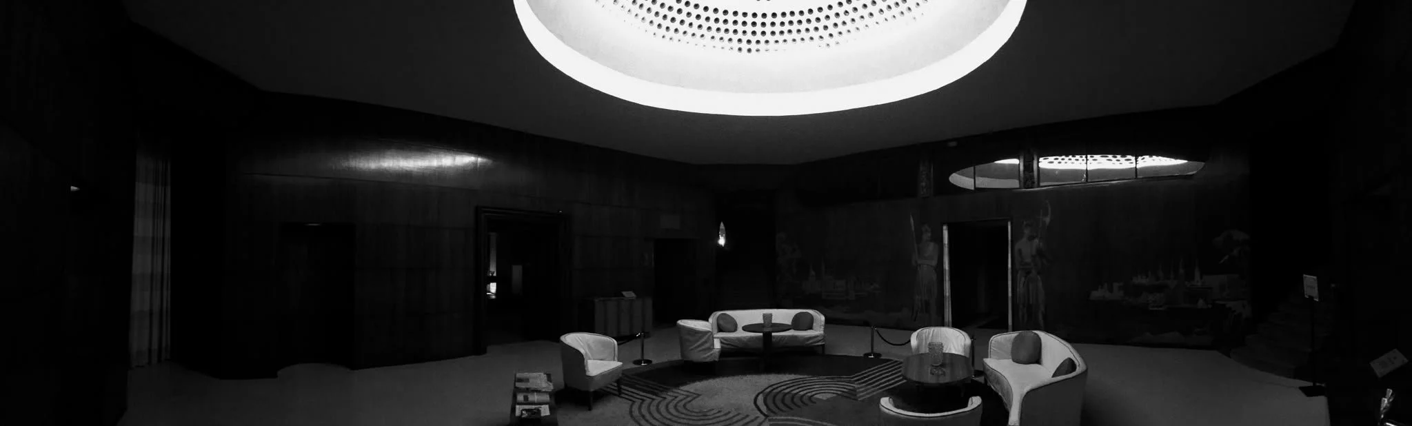 A dimly lit, modern lounge with a circular seating area and contemporary furniture, illuminated by a large circular ceiling light.