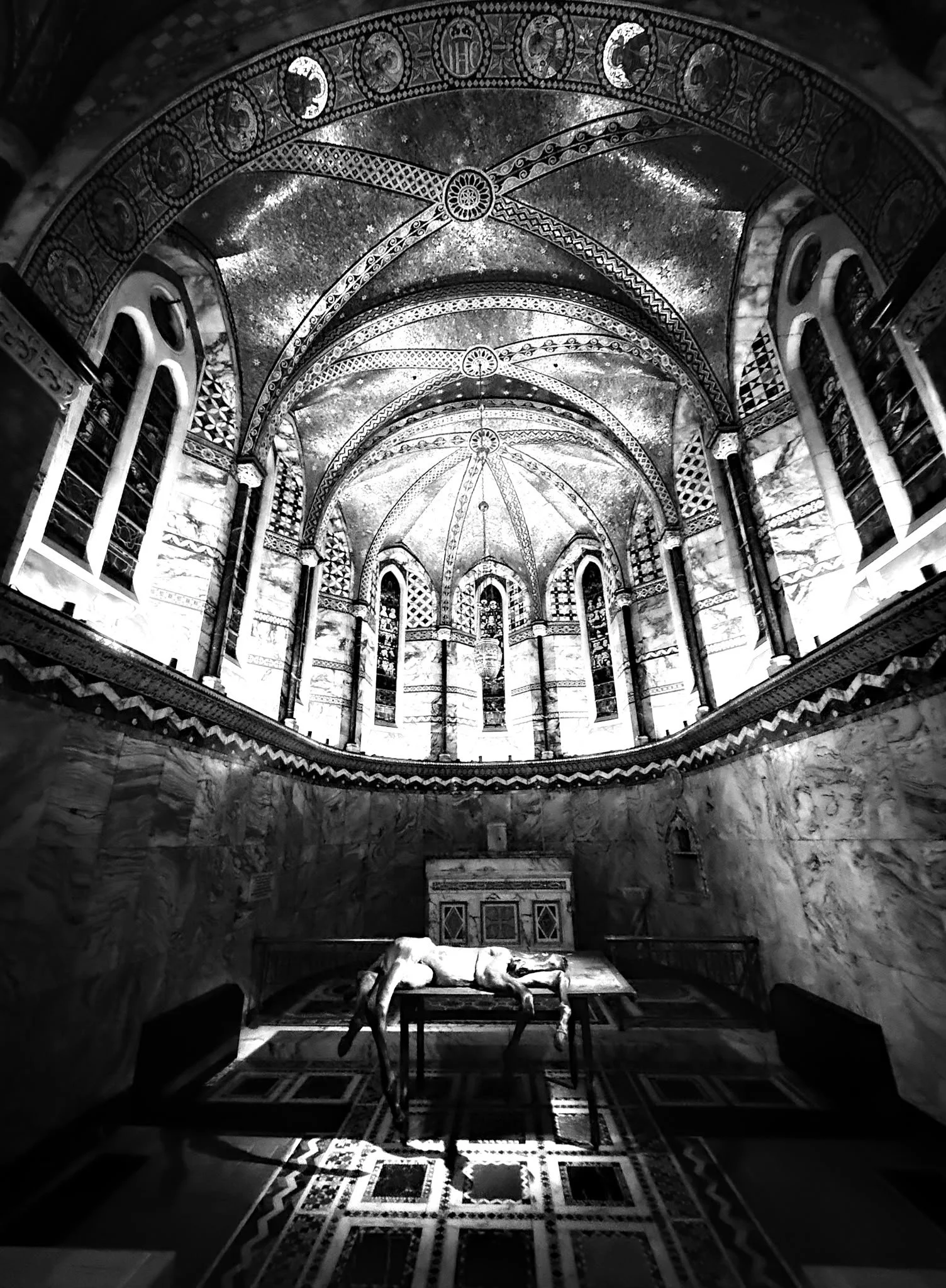 An ornate cathedral interior with high vaulted ceilings decorated with intricate patterns. A statue of an angel is lying on a table in the center of the room, with sunlight casting shadows on the patterned tiled floor.