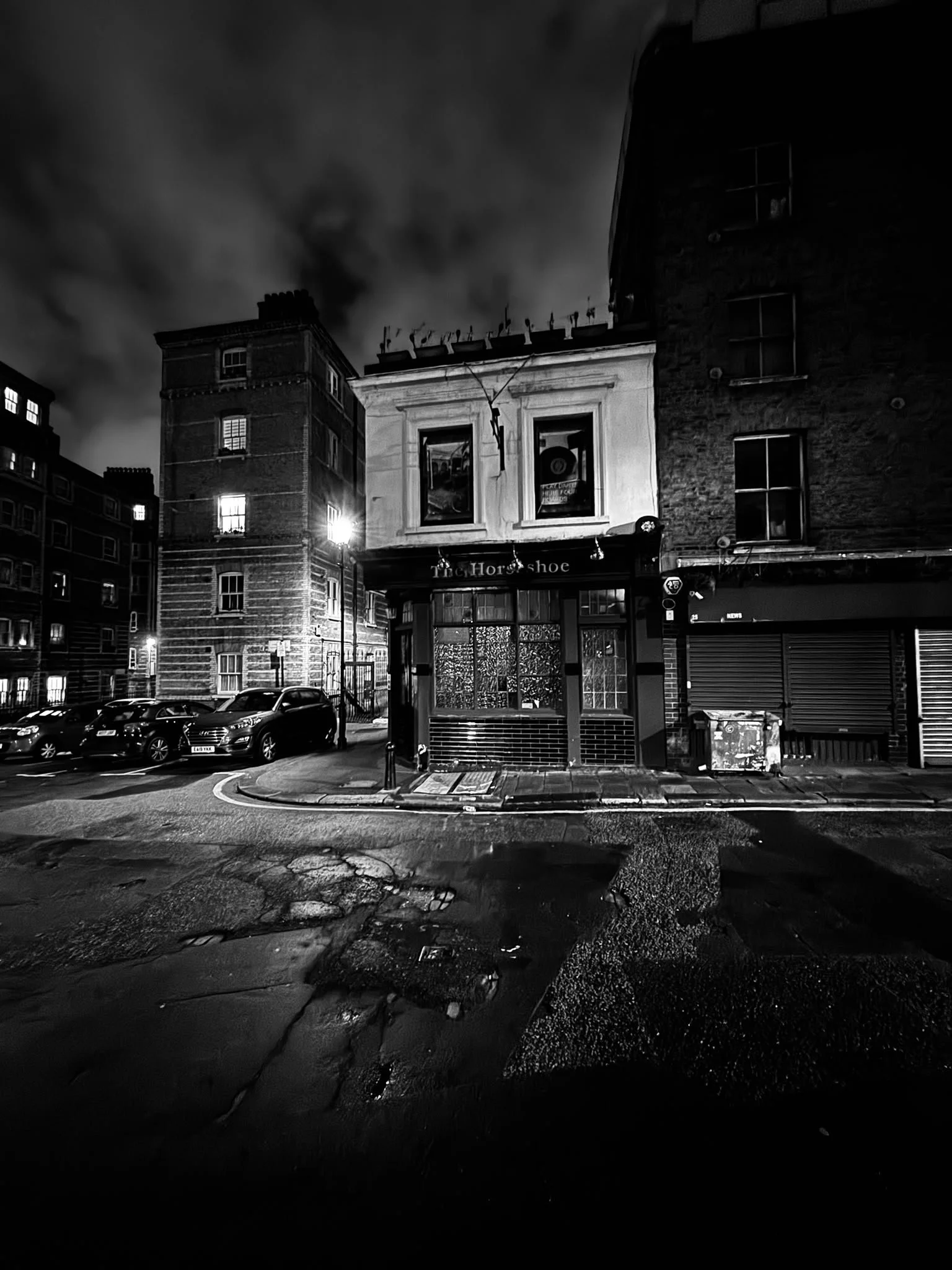 Nighttime street scene in black and white, with a row of parked cars, a small building with a sign 'The Horseshoe', and tall residential buildings in the background. The ground has cracks and puddles reflecting light.