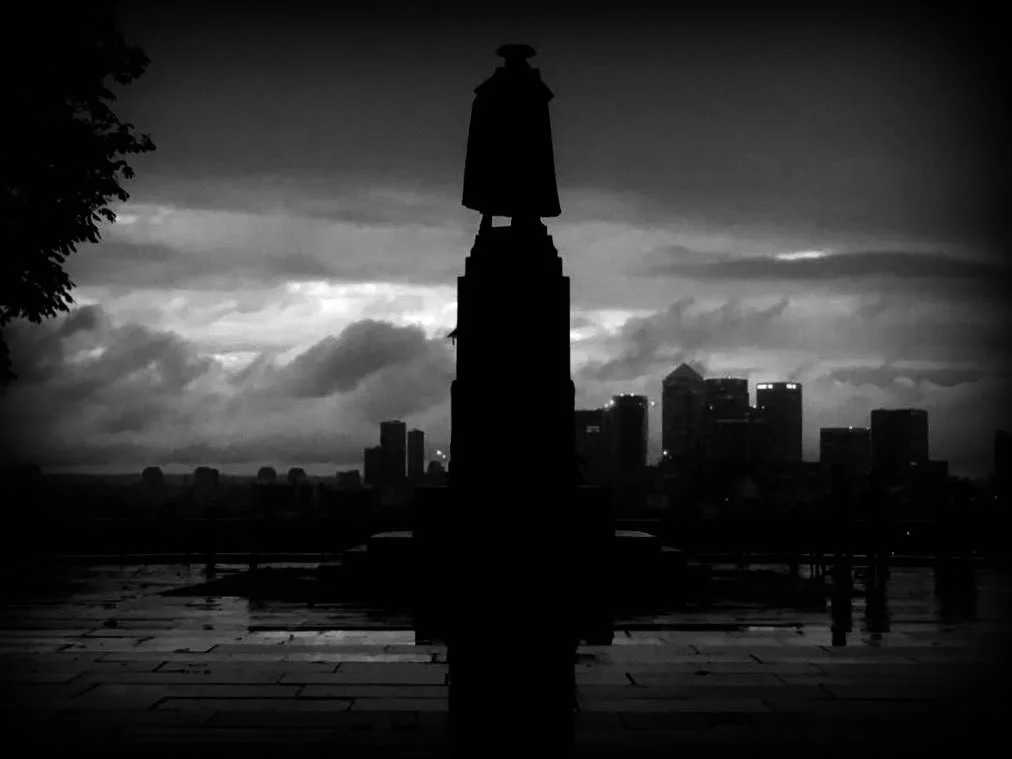 Black and white photo of a statue in front of a city skyline during cloudy weather at dusk.
