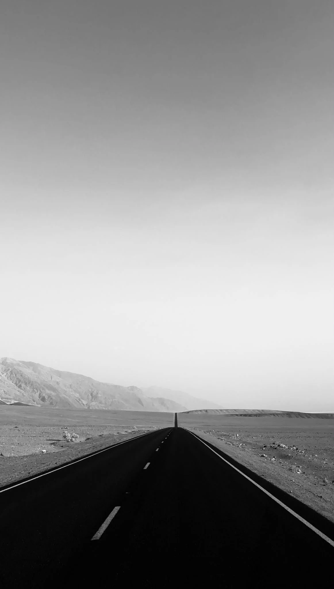 Black and white photo of a straight road stretching into the horizon, with mountains in the background and a clear sky above.