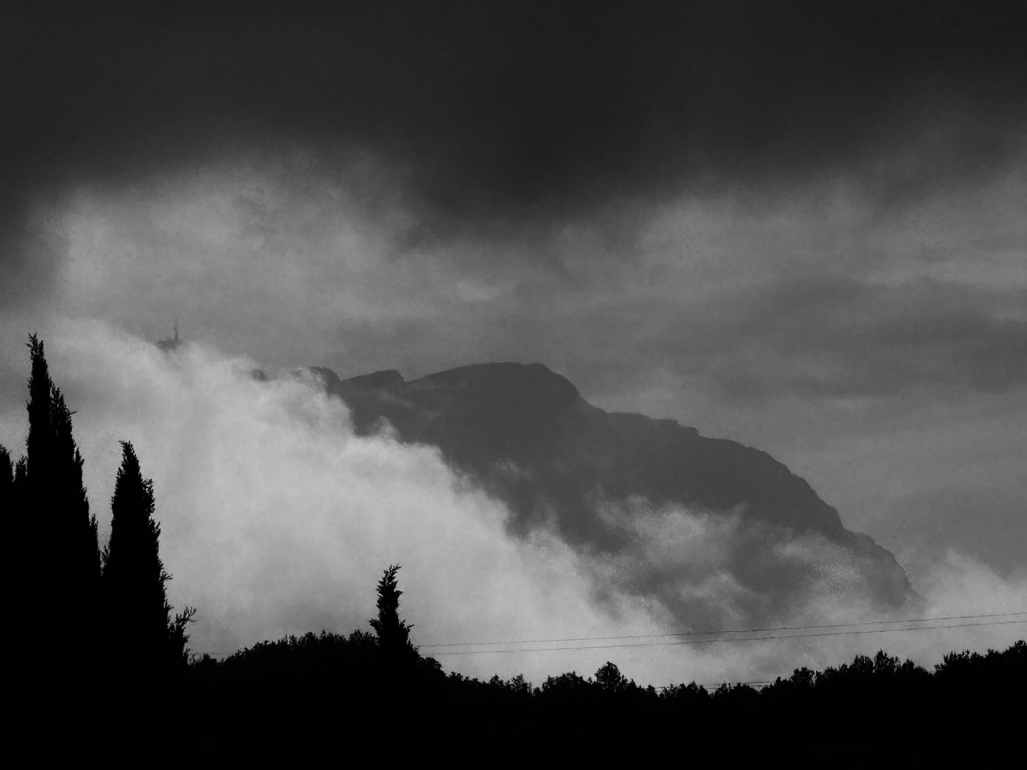 Black and white photograph of a mountain landscape with a cloudy sky, trees in the foreground, and mountains in the background.