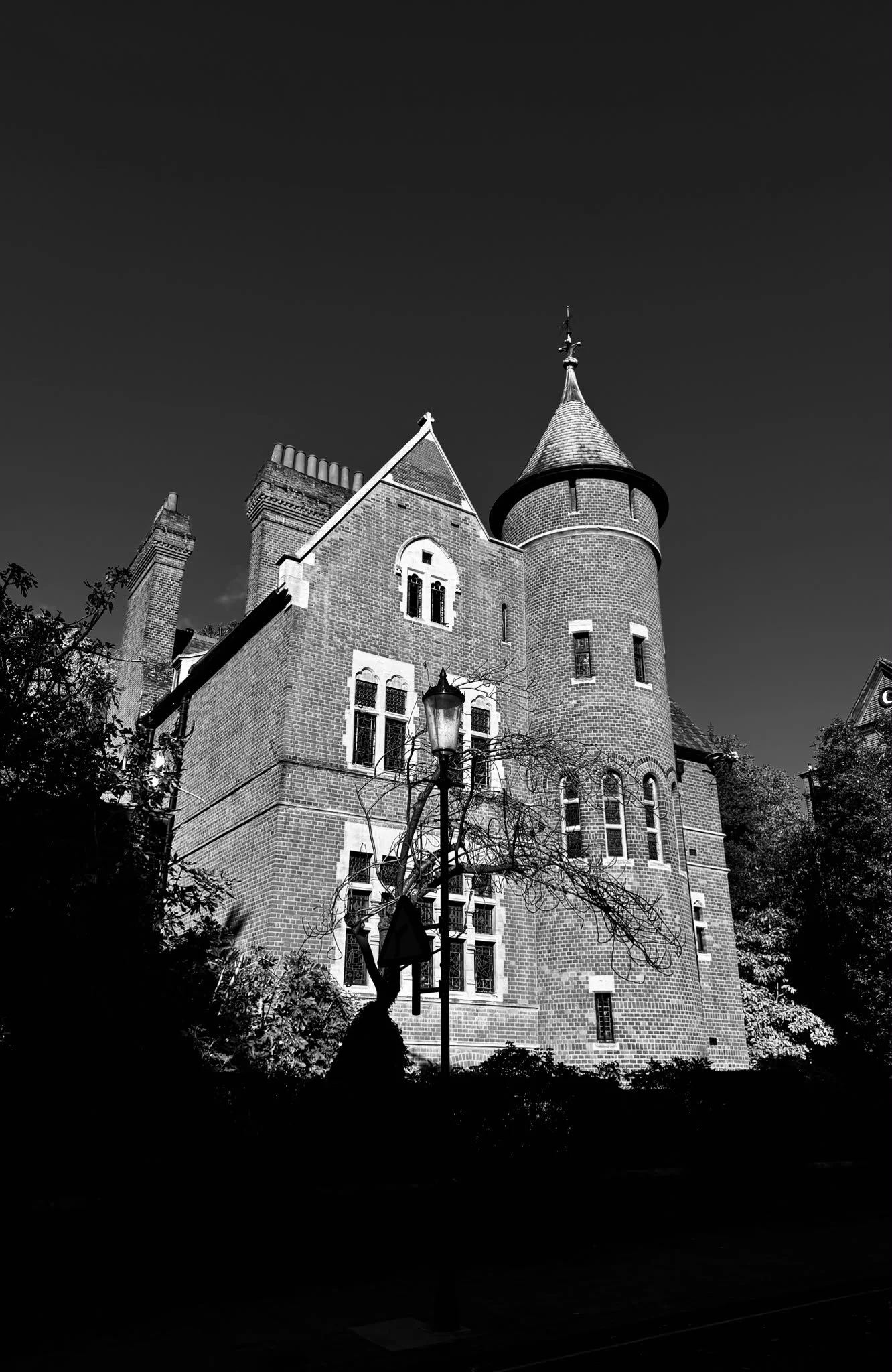 A black-and-white photo of a large, old European-style brick building with pointed roofs and a turret, surrounded by trees and a street lamp in the foreground.