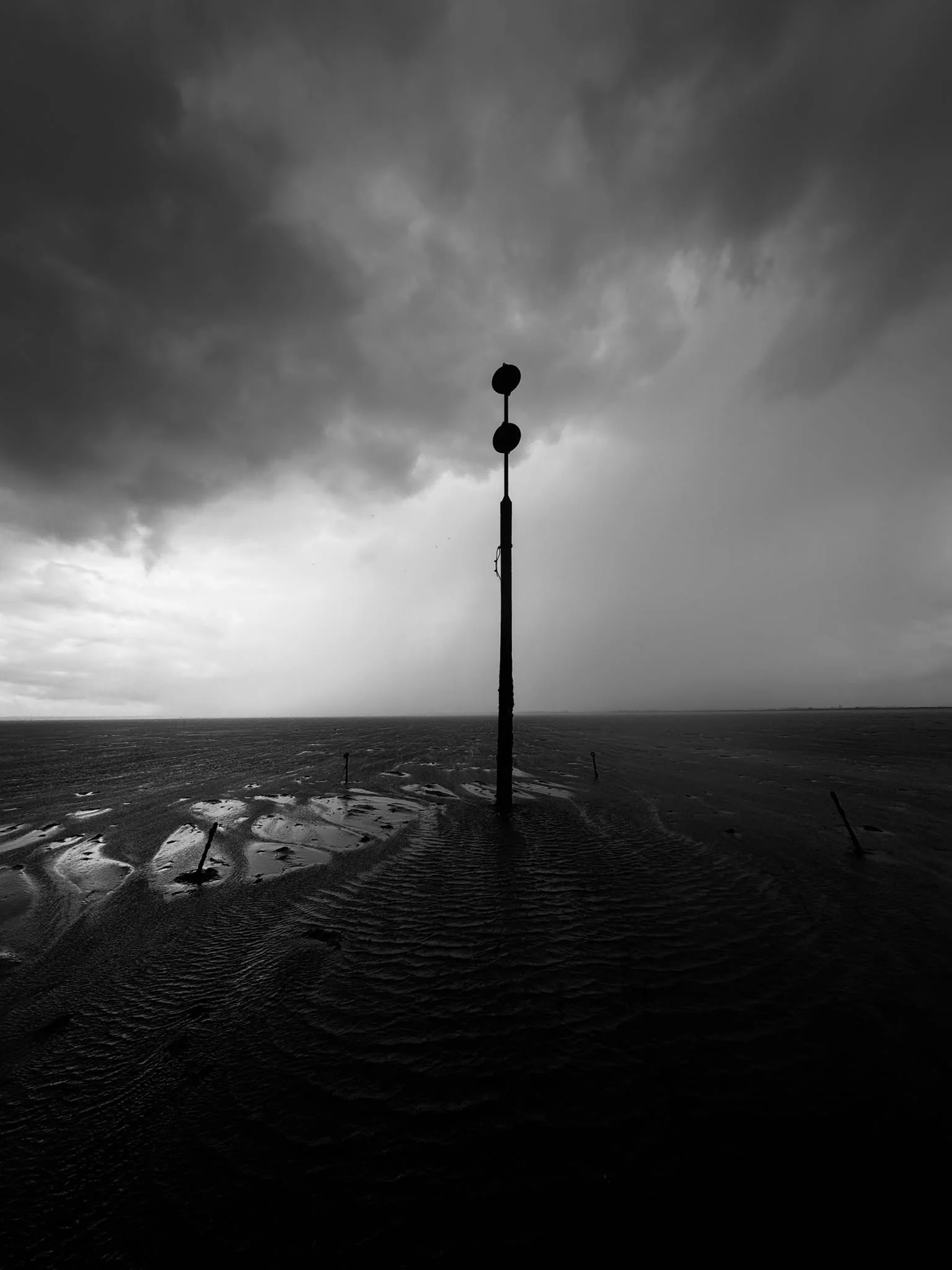 Black and white photo of a tall pole with two spherical objects on top, standing in a sandy or muddy area near water, under a cloudy sky.