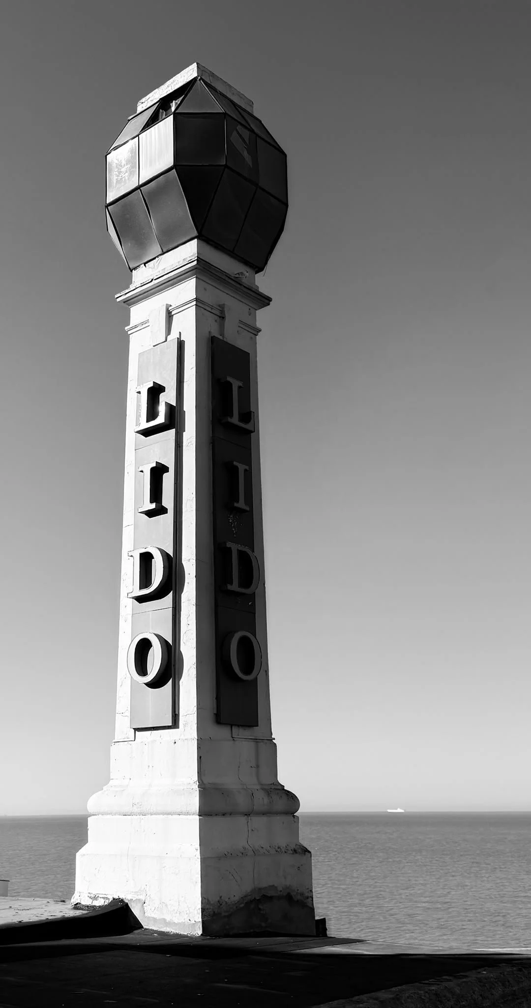 A tall, white, decorative signpost with large 3D letters spelling 'LOS ANGELES' vertically, topped with a geometric, black and metallic globe structure, against a clear sky and ocean in the background.