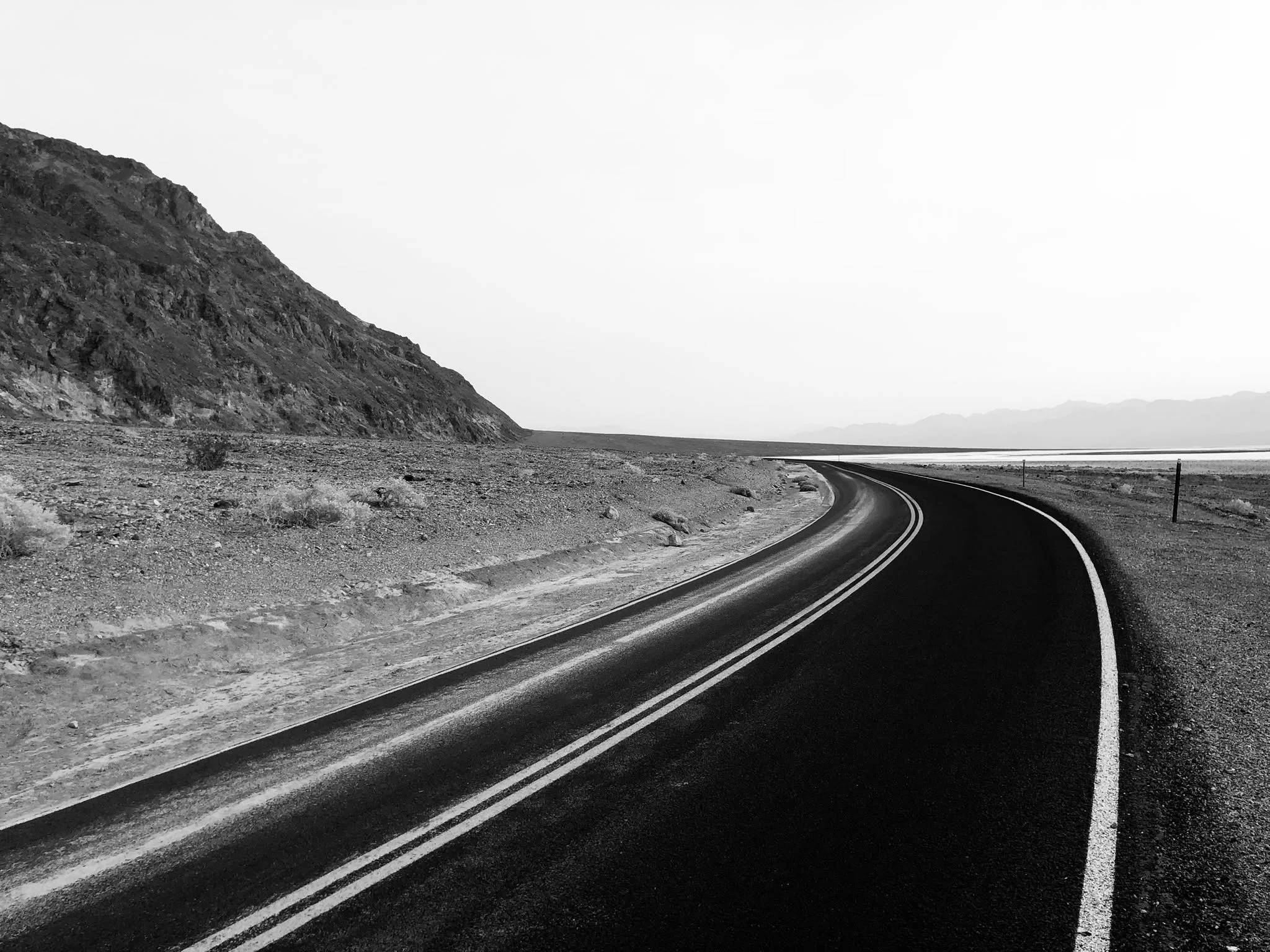 Black and white photo of a curving road through a mountainous landscape, with a rocky hillside on the left and flat land on the right.