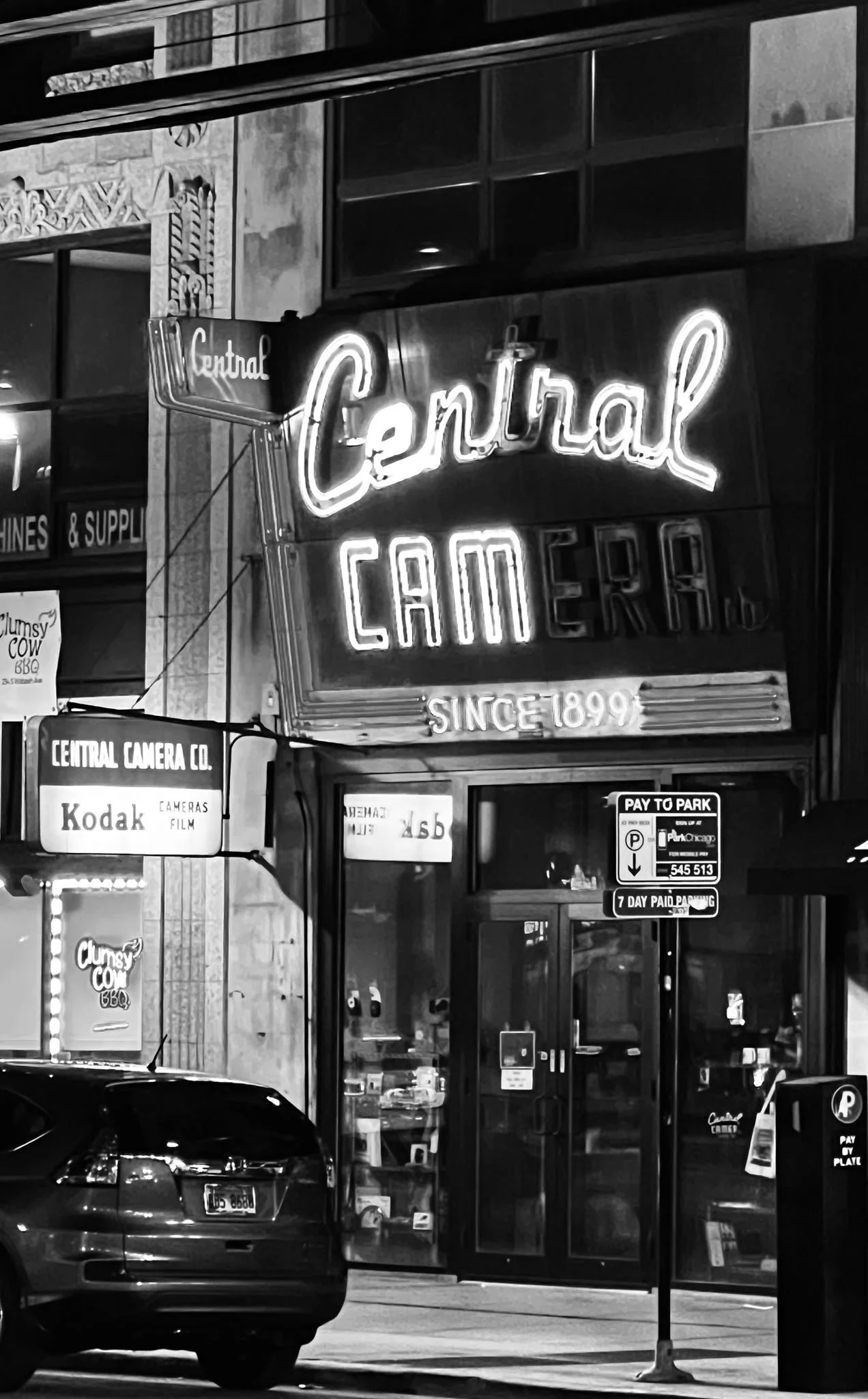 A black and white photo of a storefront with neon signs, including a large 'Central' sign and smaller signs for 'Cameras' and 'Supply.' A car is parked in front, and street signs indicate parking information.