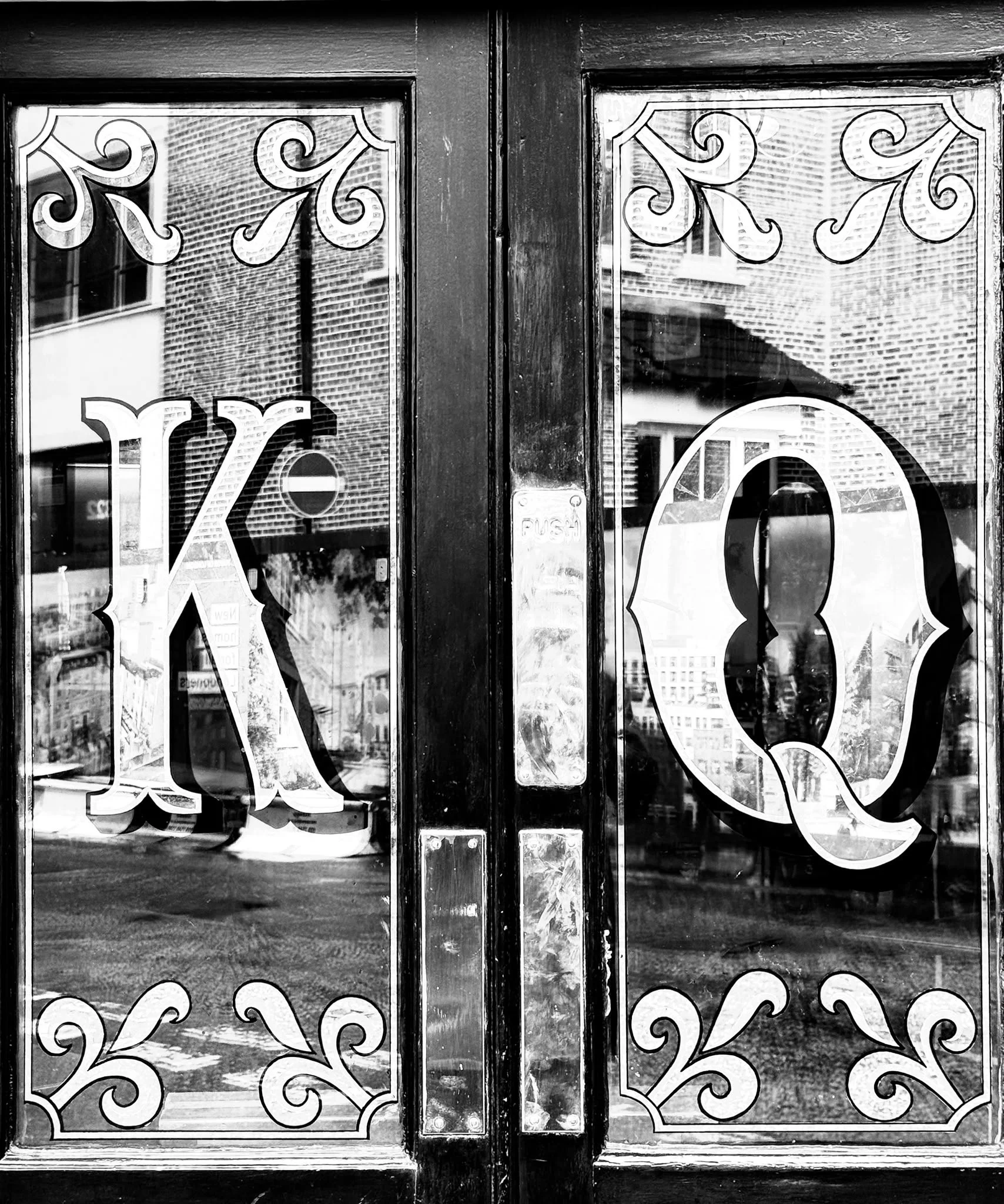 Black and white photo of a glass door with decorative white swirl patterns and the number 13. The door's reflection shows brick buildings and windows outside.
