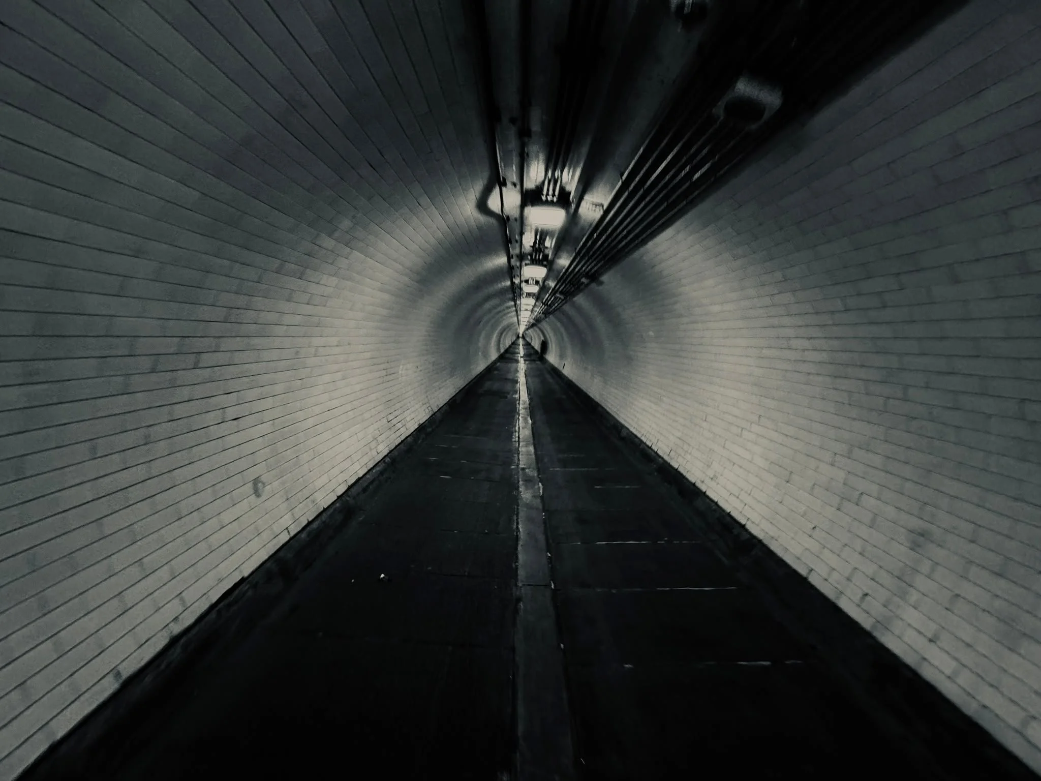 View inside a tunnel with brick walls, overhead lighting, and a straight path leading into darkness.