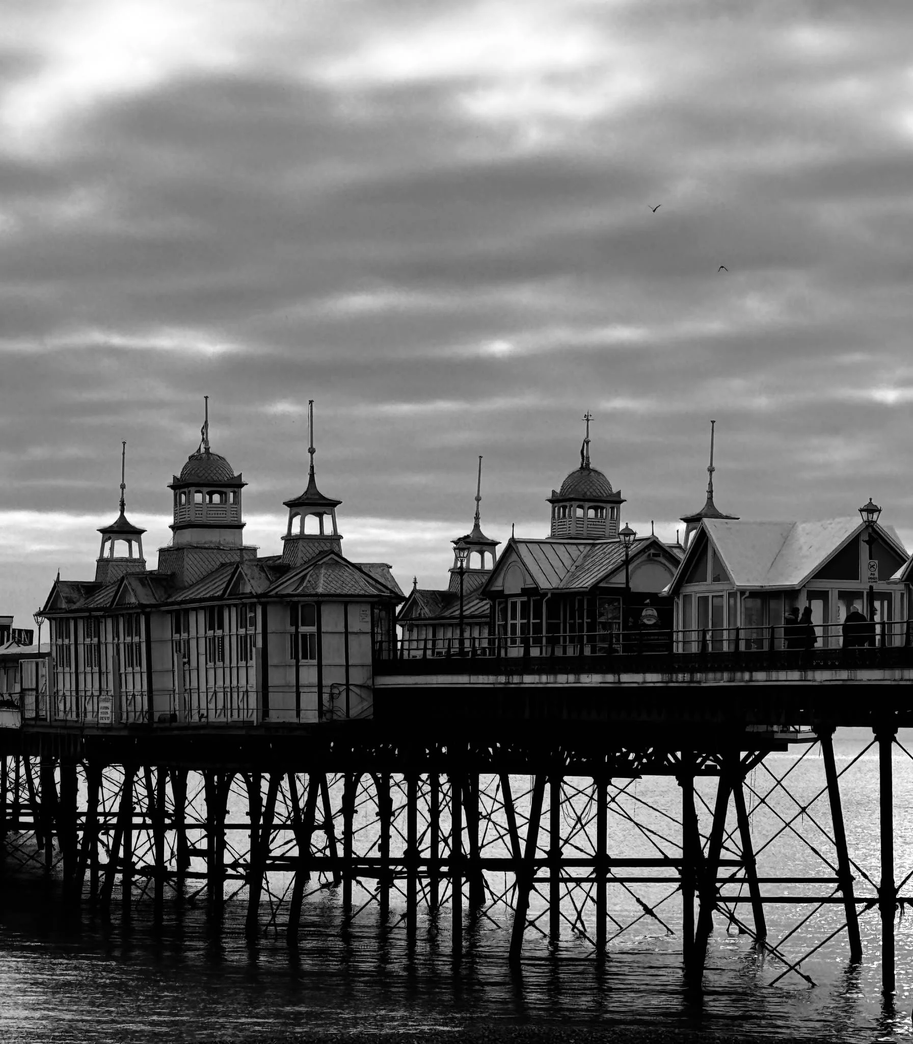 Black and white photo of a pier with ornate Victorian-style buildings featuring towers and pointed roofs, extending over water, with cloudy sky and birds flying in the background.
