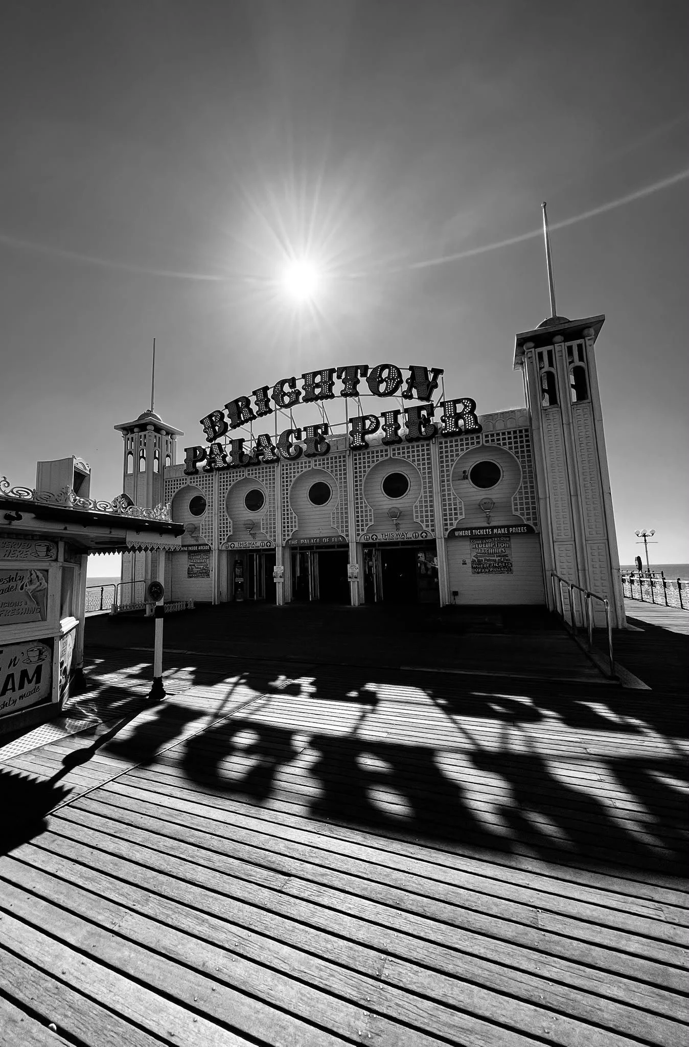 Black and white photo of the Brighton Palace Pier entrance with the sign 'Brighton Palace Pier' and decorative towers on either side, with shadows cast on a wooden boardwalk, under a bright sun.