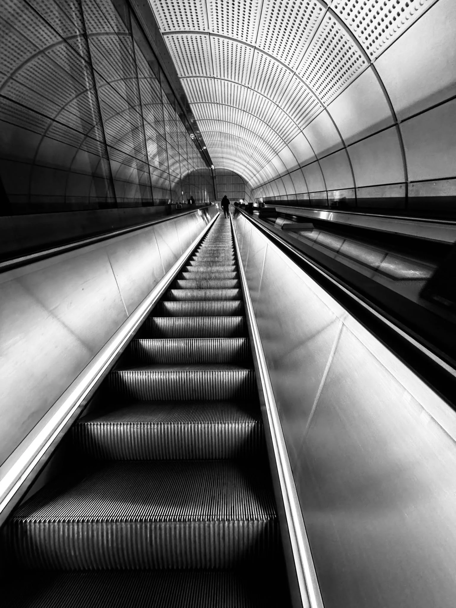 Black and white photo of an underground tunnel with an escalator in the center, leading towards a person walking in the distance.