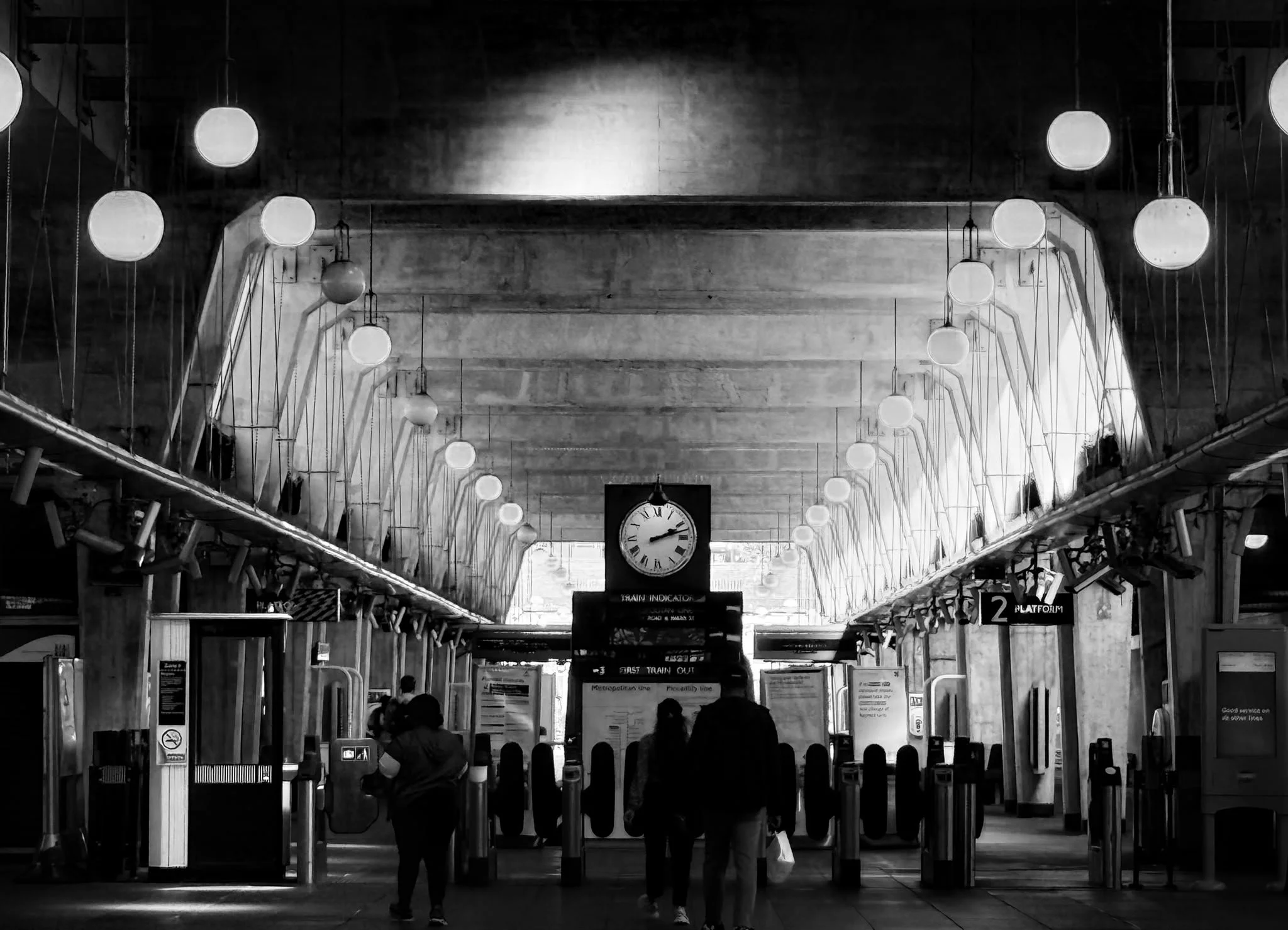 Interior of a train station with people walking and ticket barriers, large clock hanging from the ceiling, and decorative spherical lights hanging from above.