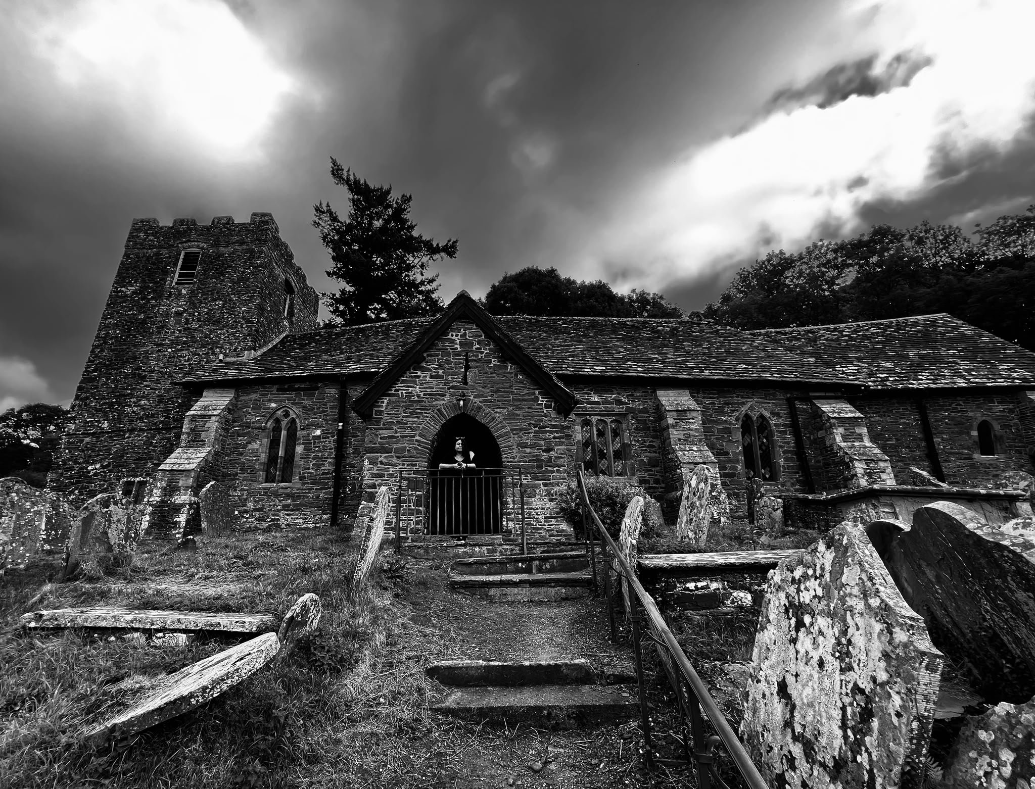Black and white photo of an old stone church with a tower, arched entrance, and gothic windows, surrounded by trees and stone steps leading to the entrance, with a woman standing on the porch.